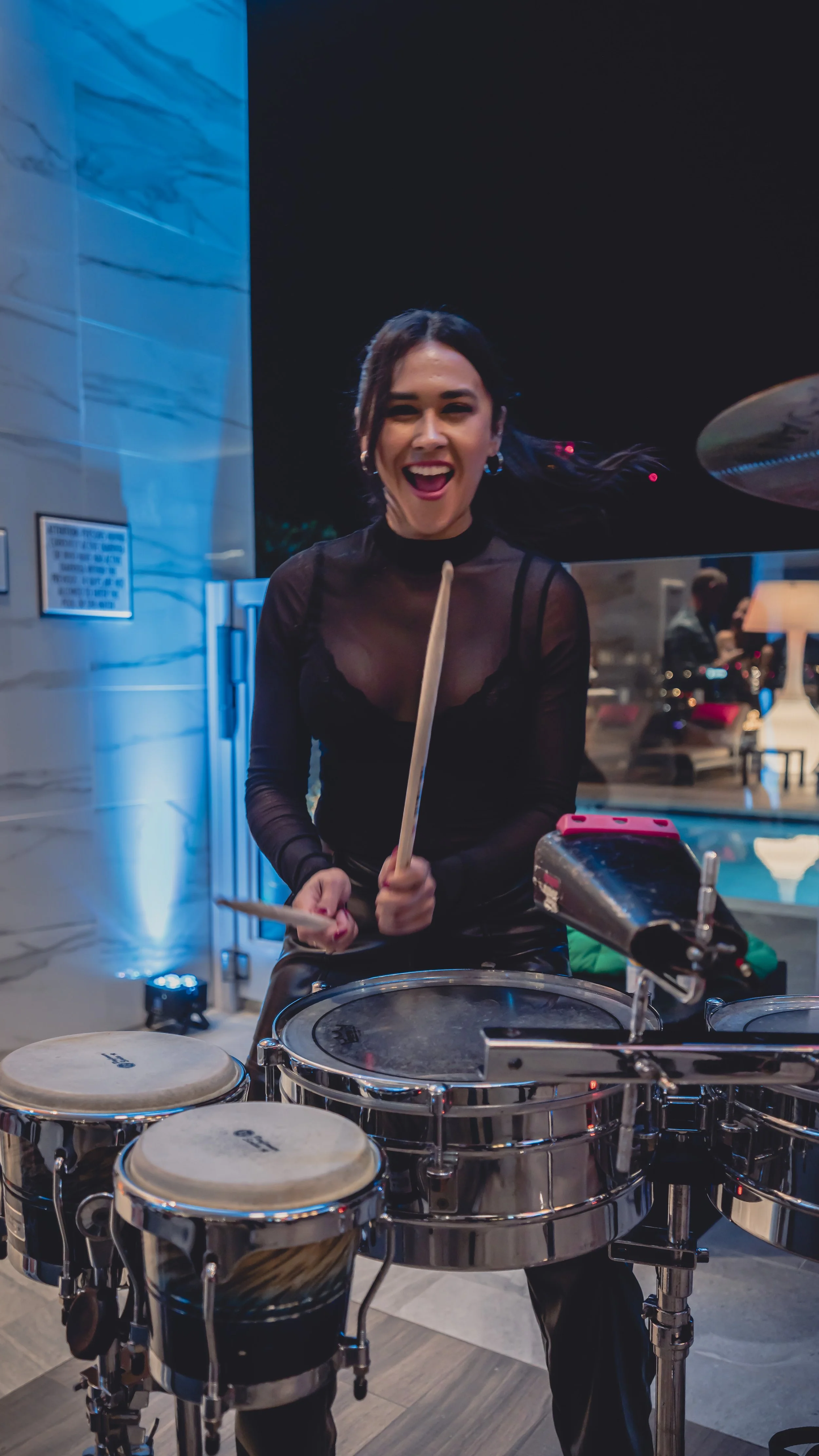 A woman smiling and playing electronic drums at an indoor event with blue accent lighting.