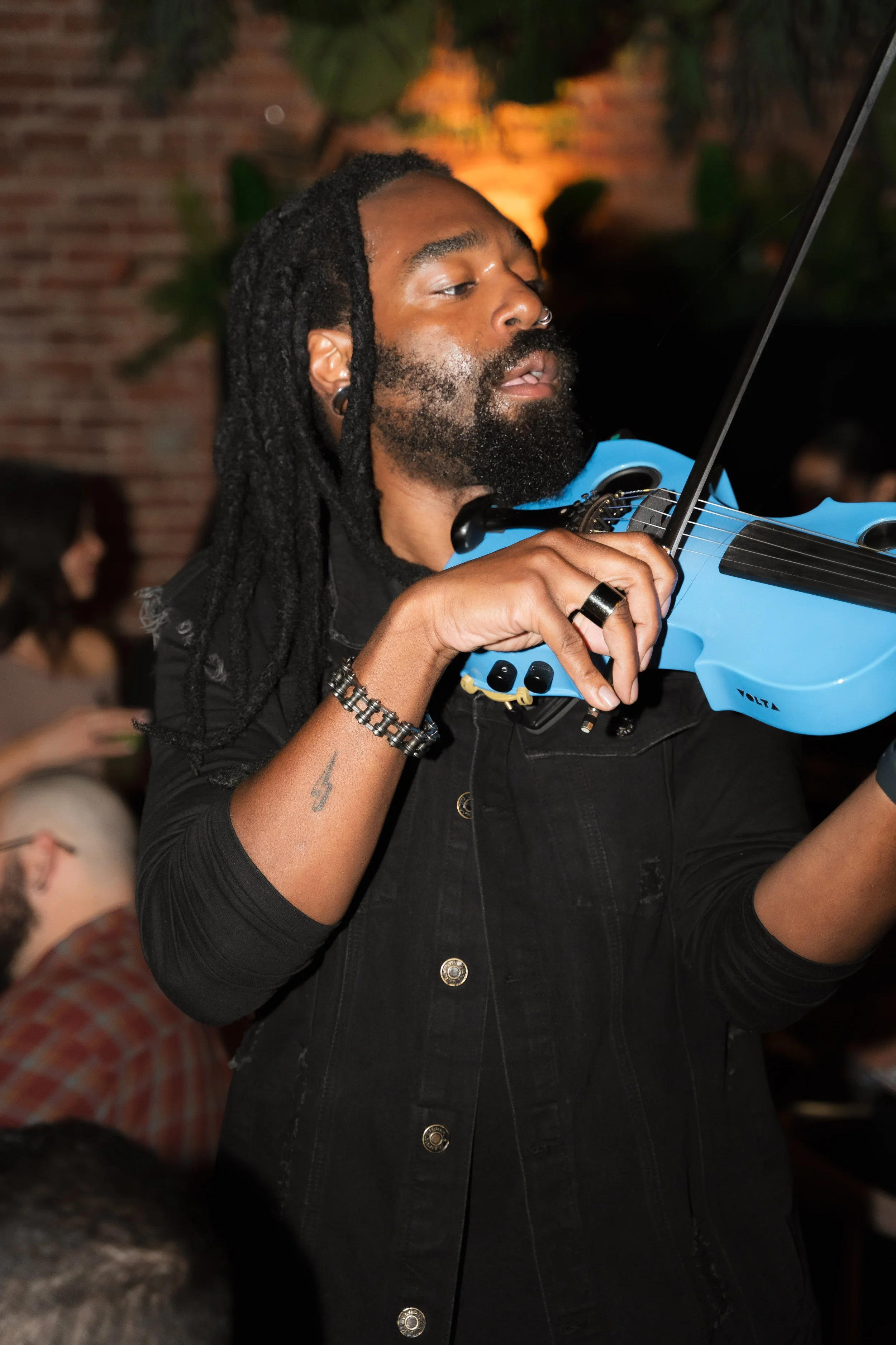 A man playing a blue electric violin at a social gathering, with a brick wall and blurred people in the background.