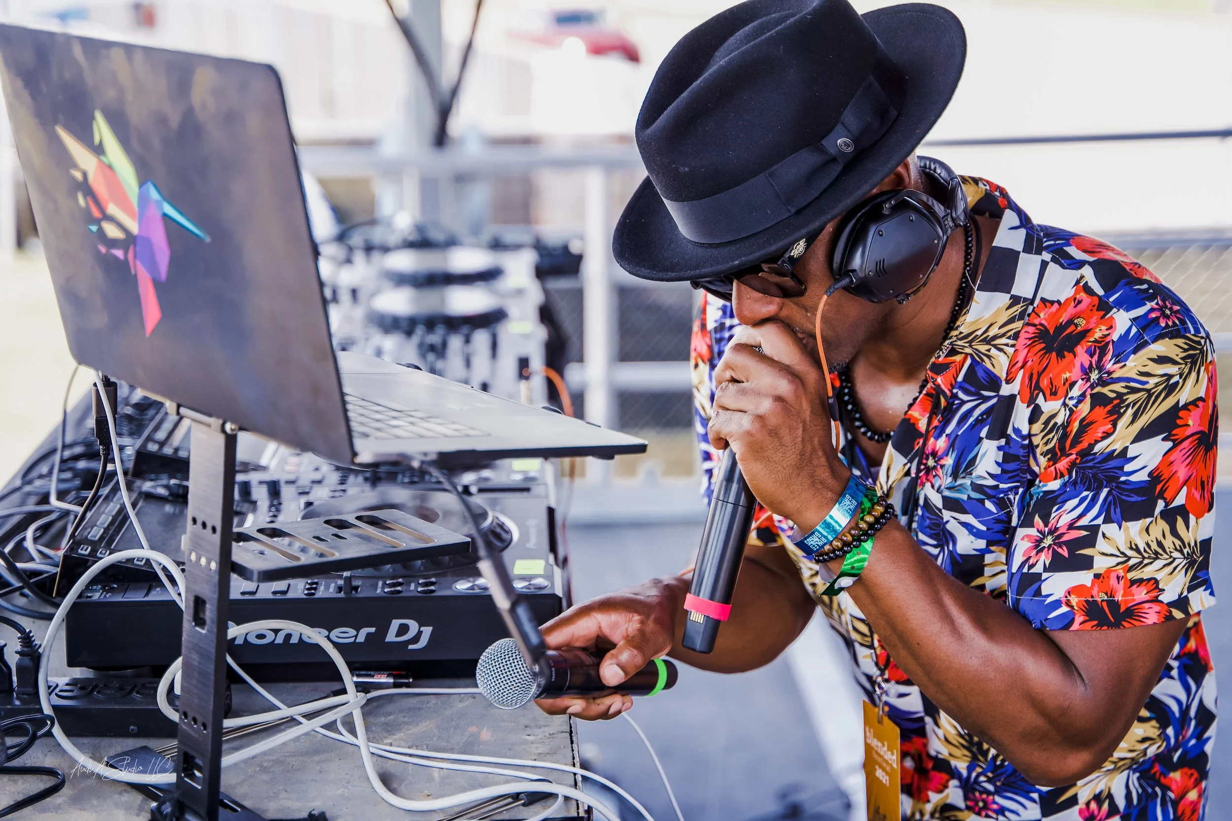 A DJ wearing a black hat, sunglasses, and colorful tropical shirt, holding a microphone near DJ equipment and a laptop in an outdoor setting.