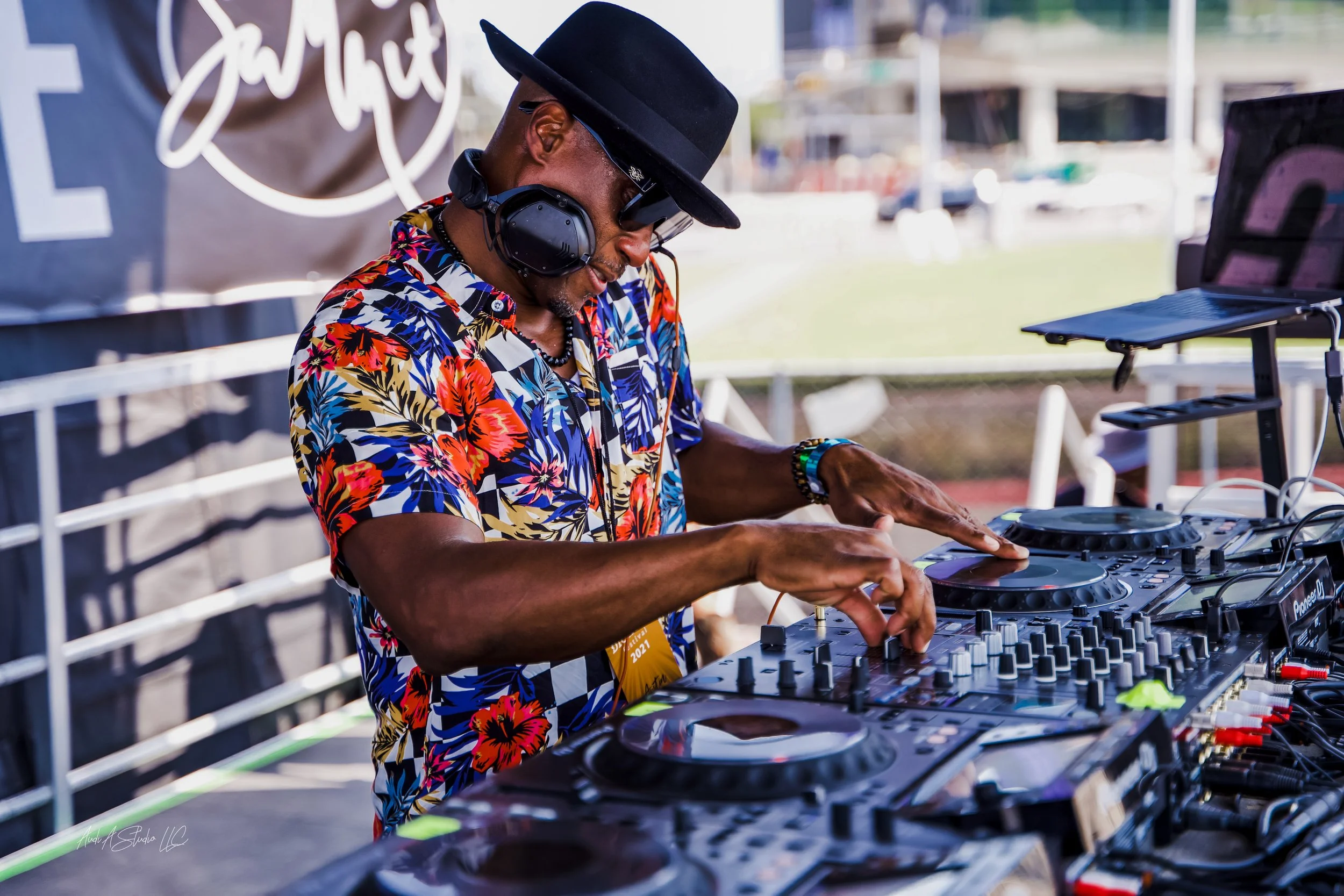 A DJ wearing a colorful floral shirt, black hat, sunglasses, and headphones, operating DJ equipment outdoors.