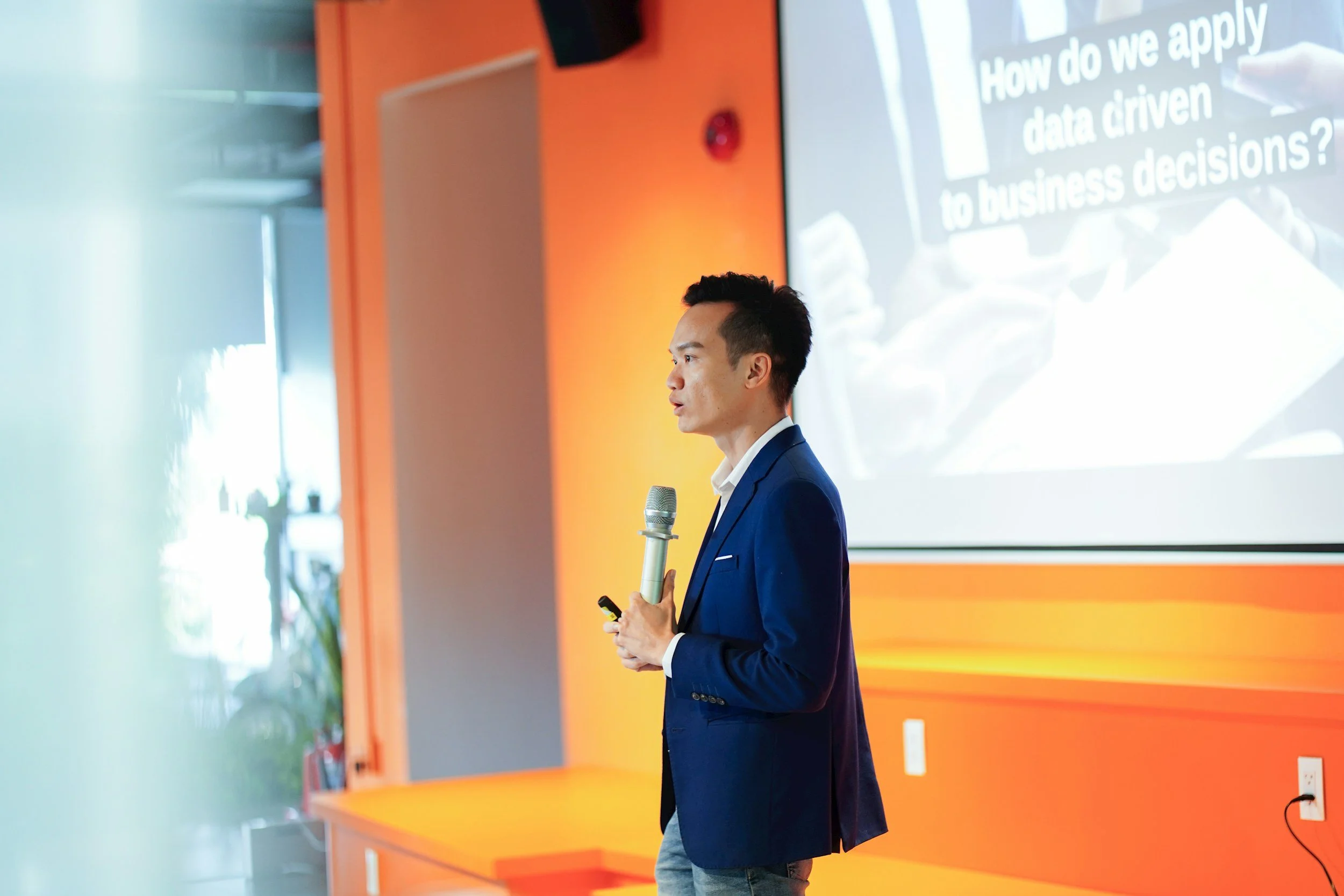 A man in a blue suit giving a presentation in front of a large screen with text about applying data-driven commercial real estate business decisions.