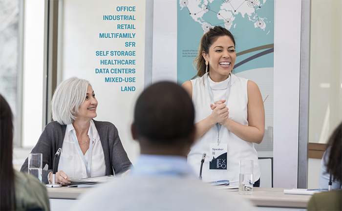 A woman giving a commercial real estate topic presentation at a seminar, with an audience seated in front of her, and a list of different real estate property types on a banner behind her.