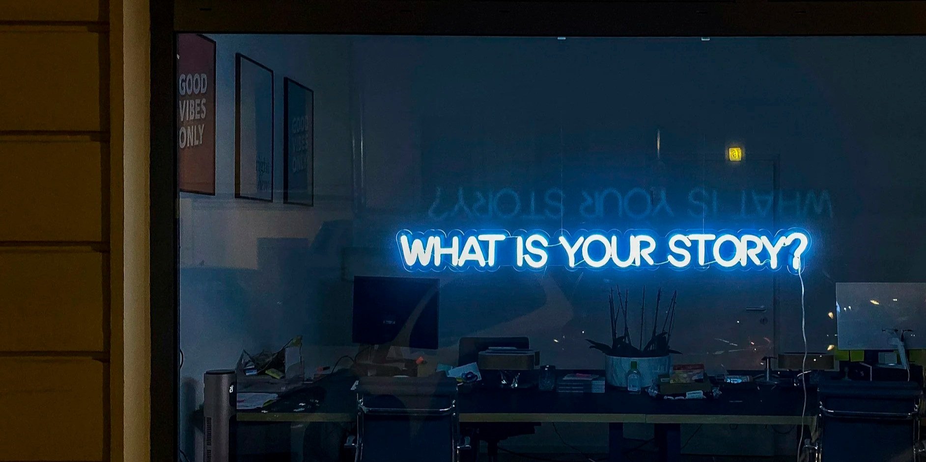 Neon sign in a window displaying the question "WHAT IS YOUR STORY?" with a reflection of the text on the glass.
