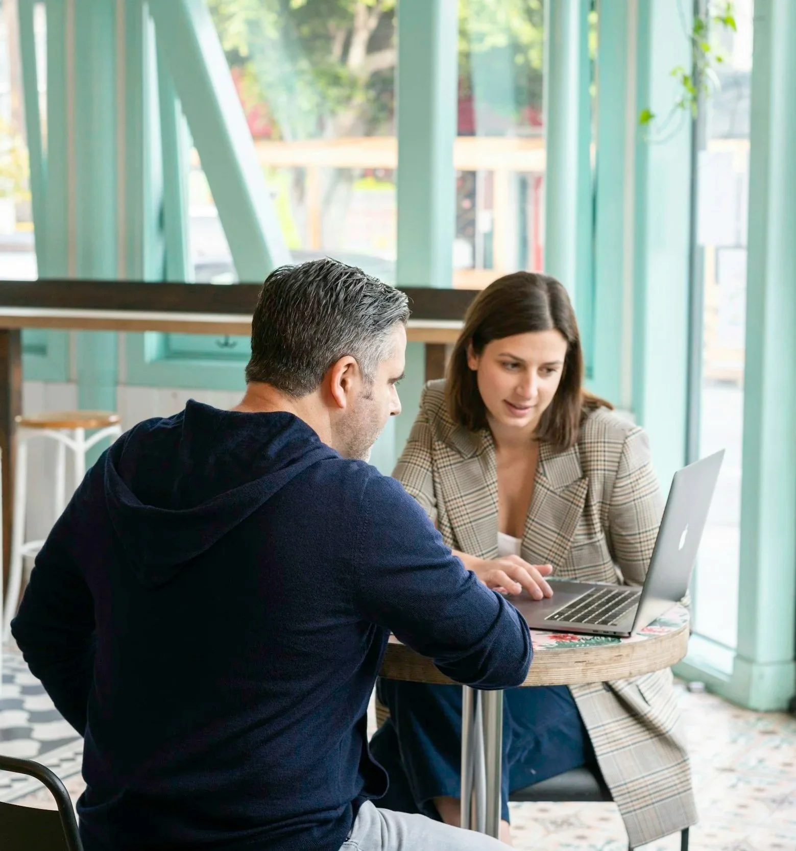 Two people, a man and a woman, sitting at a round table in a brightly lit cafe, looking at a laptop together.