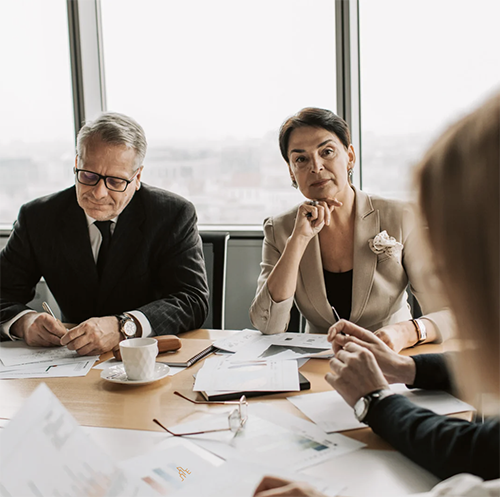 A business meeting with three people, two women and one man, seated at a table with documents, glasses, and a cup, in an office with large windows.