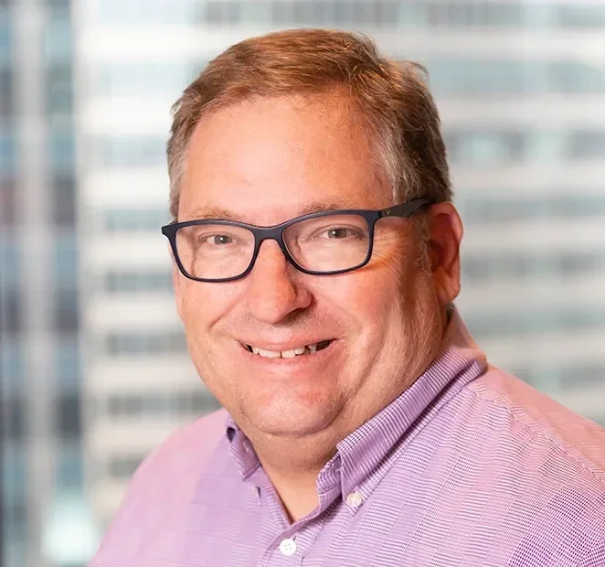 A corporate headshot of a man with glasses, short light brown hair, wearing a purple collared shirt, smiling, with a cityscape background.