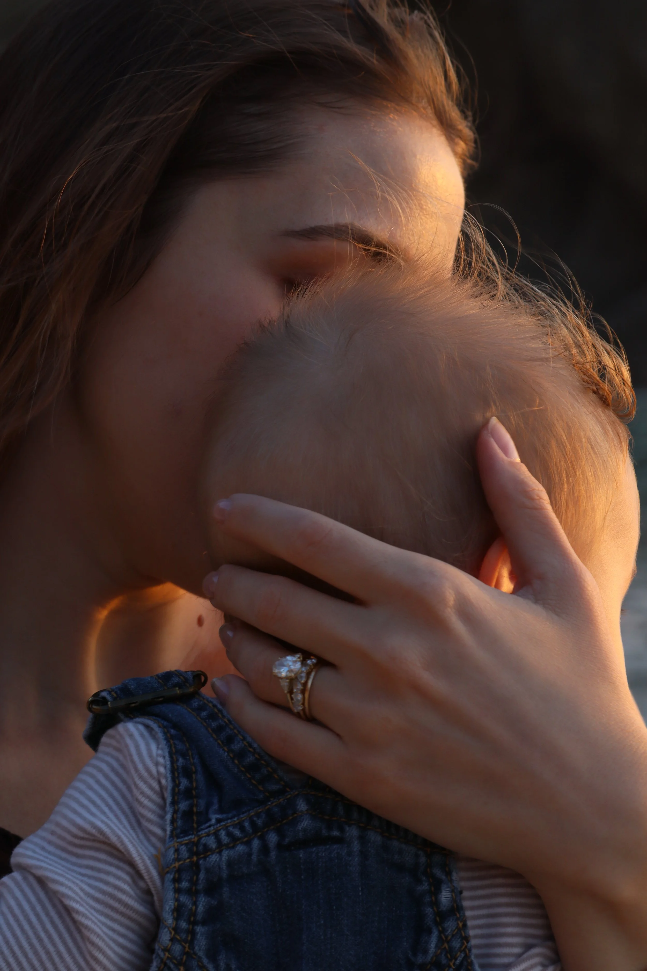 A woman with brown hair is hugging a person with light-colored hair, with her hand resting on the person's head. The woman is wearing rings and a striped top, and they are outdoors during sunset.