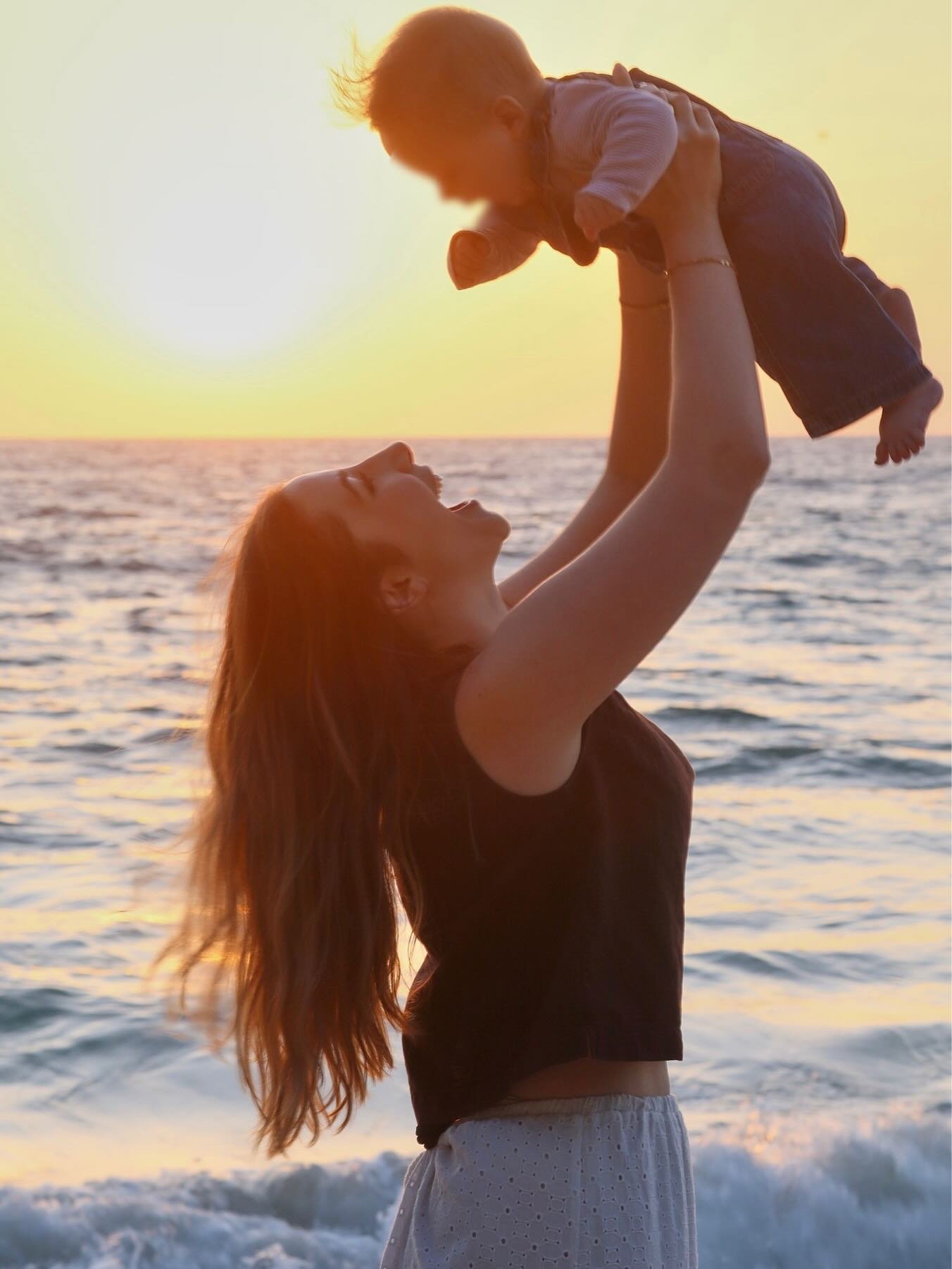 A woman with long red hair lifting a young child in the air by the ocean during sunset, both smiling and enjoying the moment.