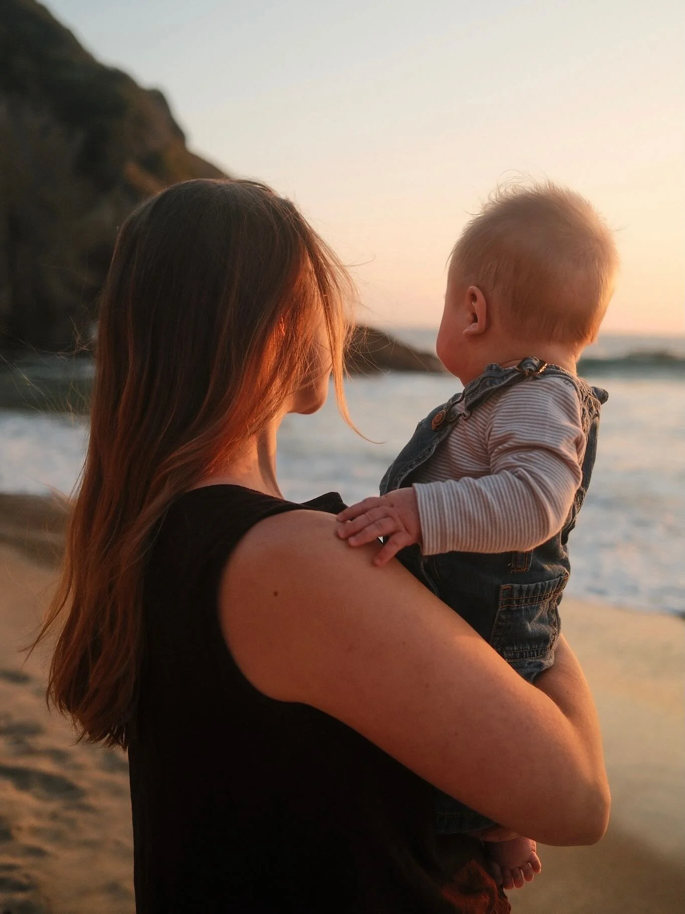 A woman with long brown hair holding a young blond-haired boy in her arms on the beach during sunset.