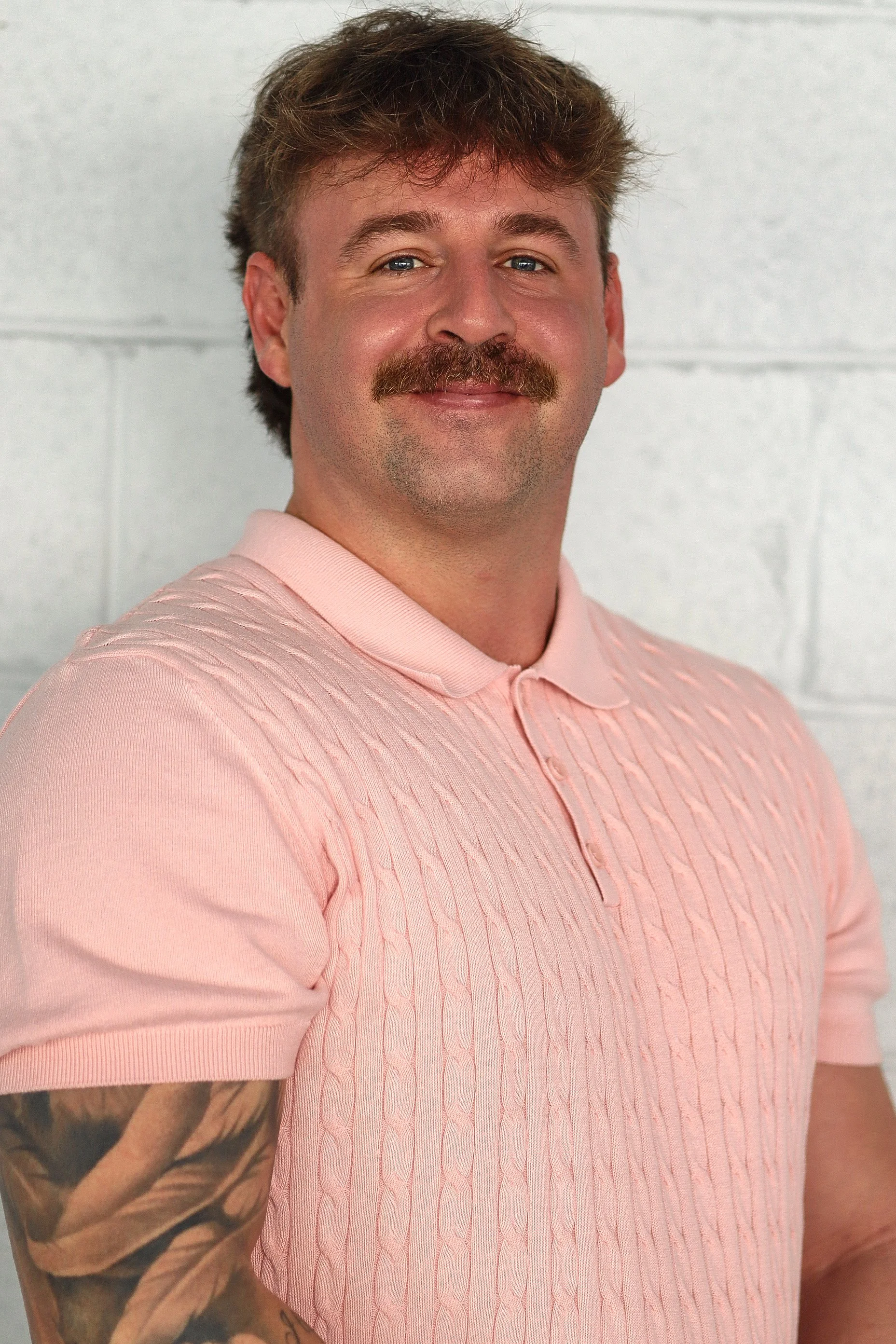 A man with brown hair, a mustache, and a beard wearing a light pink short sleeve knit polo shirt, smiling slightly and standing against a plain light-colored wall.
