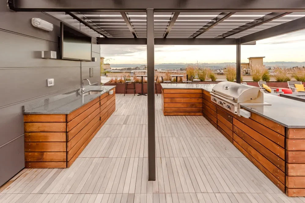 Rooftop outdoor kitchen with a grill, sink, TV, and seating area, overlooking a city skyline and body of water.