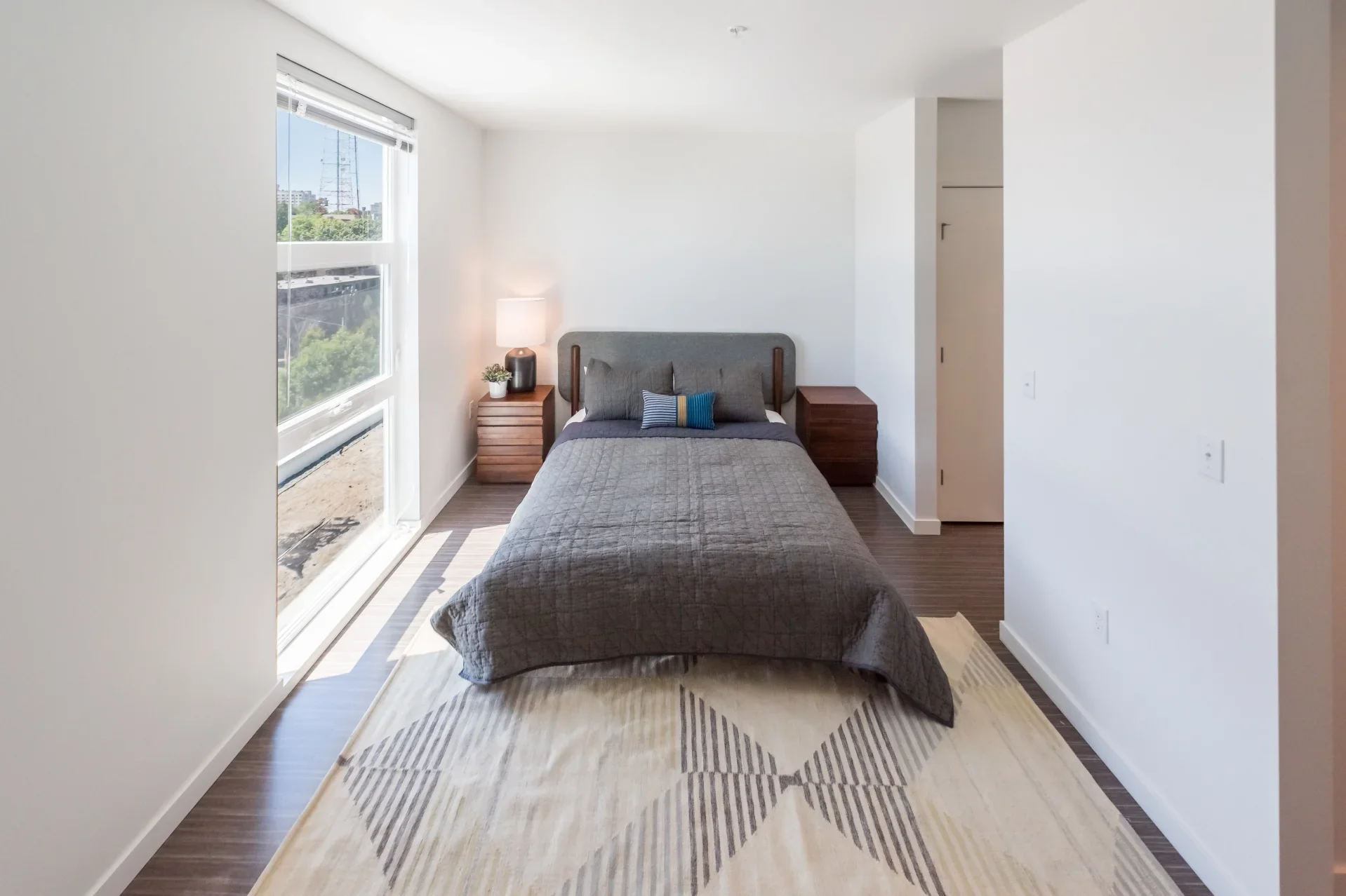 A modern bedroom with white walls and a large window. The bed has a gray headboard, gray quilt, and decorative pillows. There are wooden nightstands on each side of the bed, with a lamp and a small plant on one. A landscape painting is partially visi
