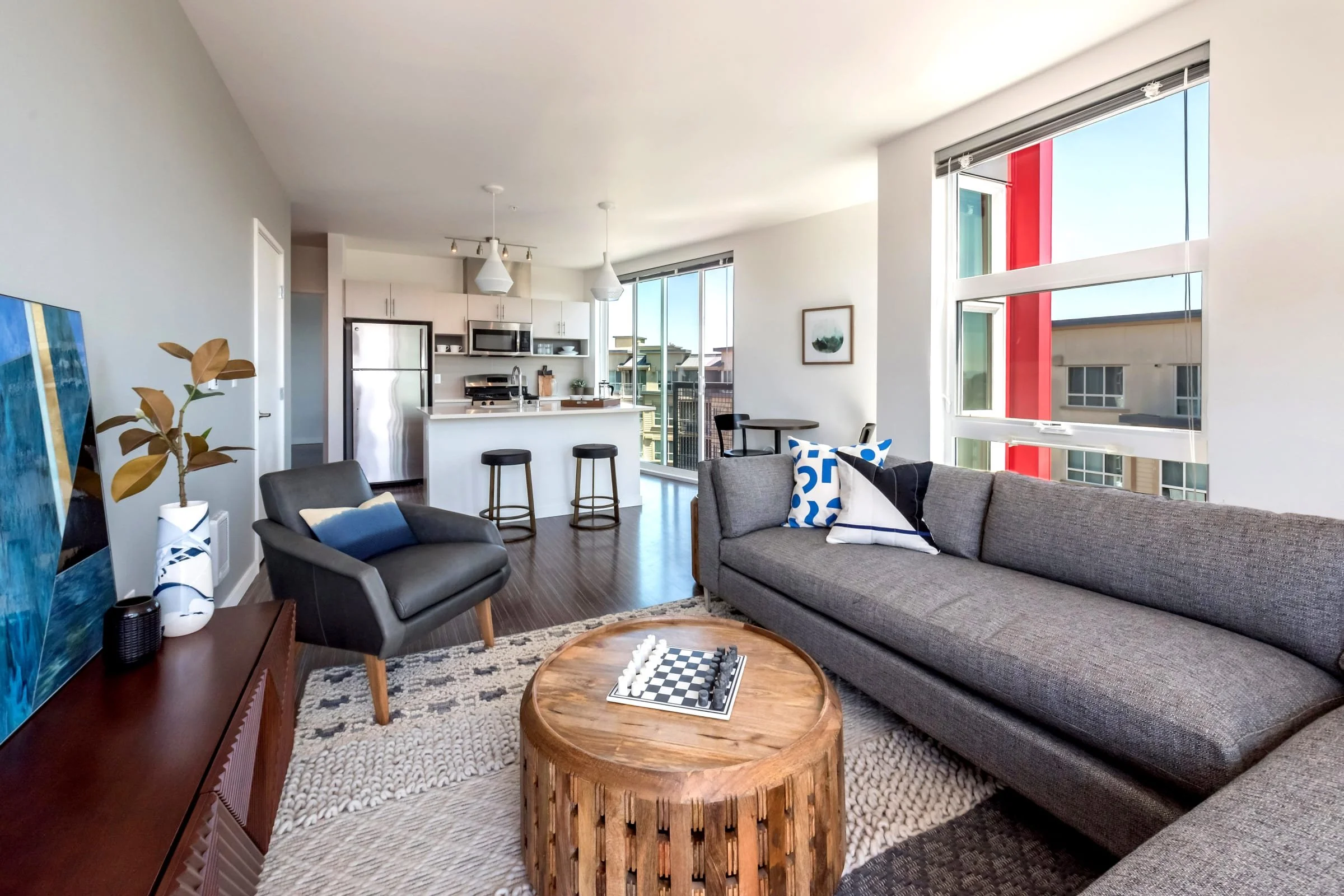Interior of a modern apartment living room with large windows, gray sofa with patterned pillows, overstuffed chair, round wooden coffee table with a chessboard, television on a sideboard, potted plant, and open kitchen with bar stools and pendant lig