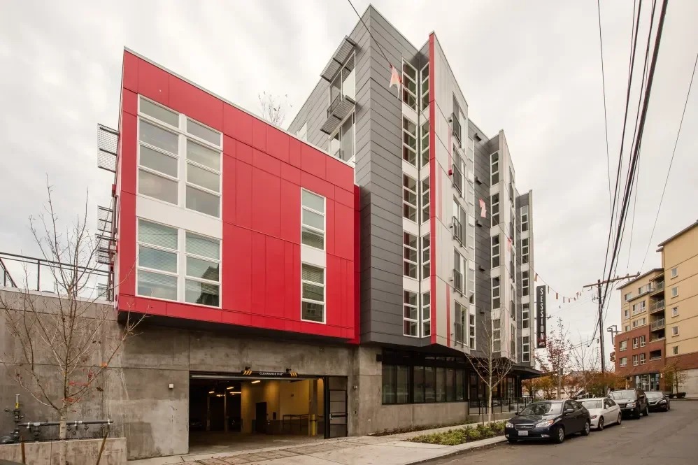 Modern multi-story apartment building with red and gray exterior, large windows, and a ground-floor parking garage, located on a city street with parked cars and leafless trees.