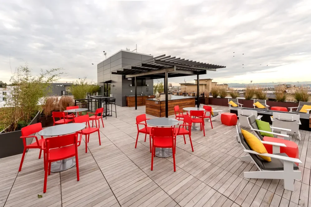 A rooftop deck with red and gray chairs, tables, and lounge chairs, some with yellow and green cushions, and a bar area with a shaded structure, plants, and a city skyline in the background.