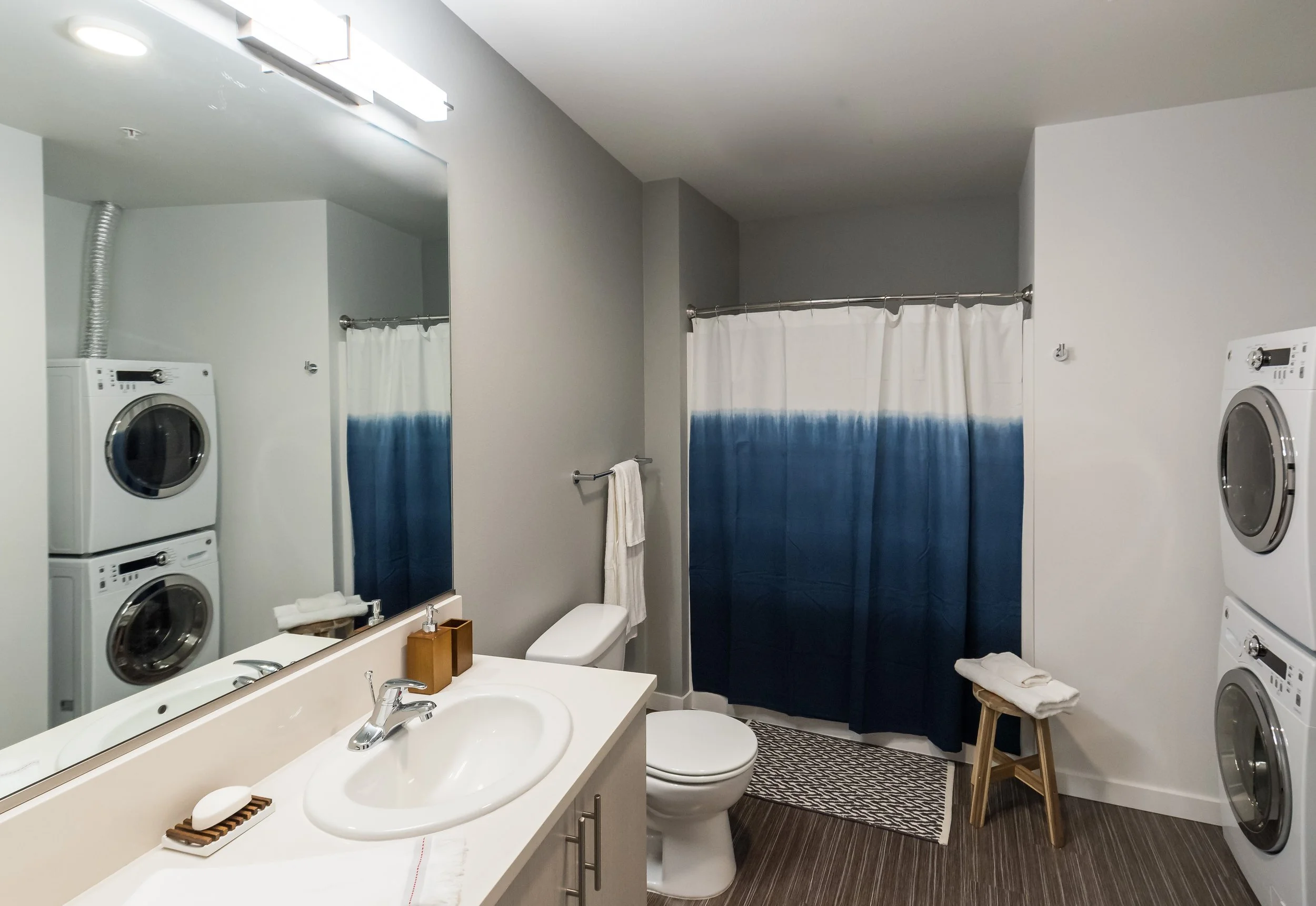 A bathroom with a stacked washer and dryer, a white toilet, a white vanity with a sink, a mirror, a blue and white gradient shower curtain, a towel rack with a towel, and a small table with towels.