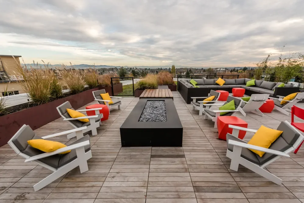 Rooftop patio with modern outdoor furniture, including white lounge chairs with yellow and green pillows, red and gray side tables, and a low fireplace, overlooking a cityscape with cloudy sky.