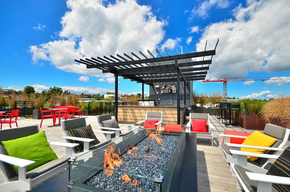 Rooftop terrace with modern seating, firepit table, bar area, outdoor dining tables, and city skyline view, under blue sky with clouds.