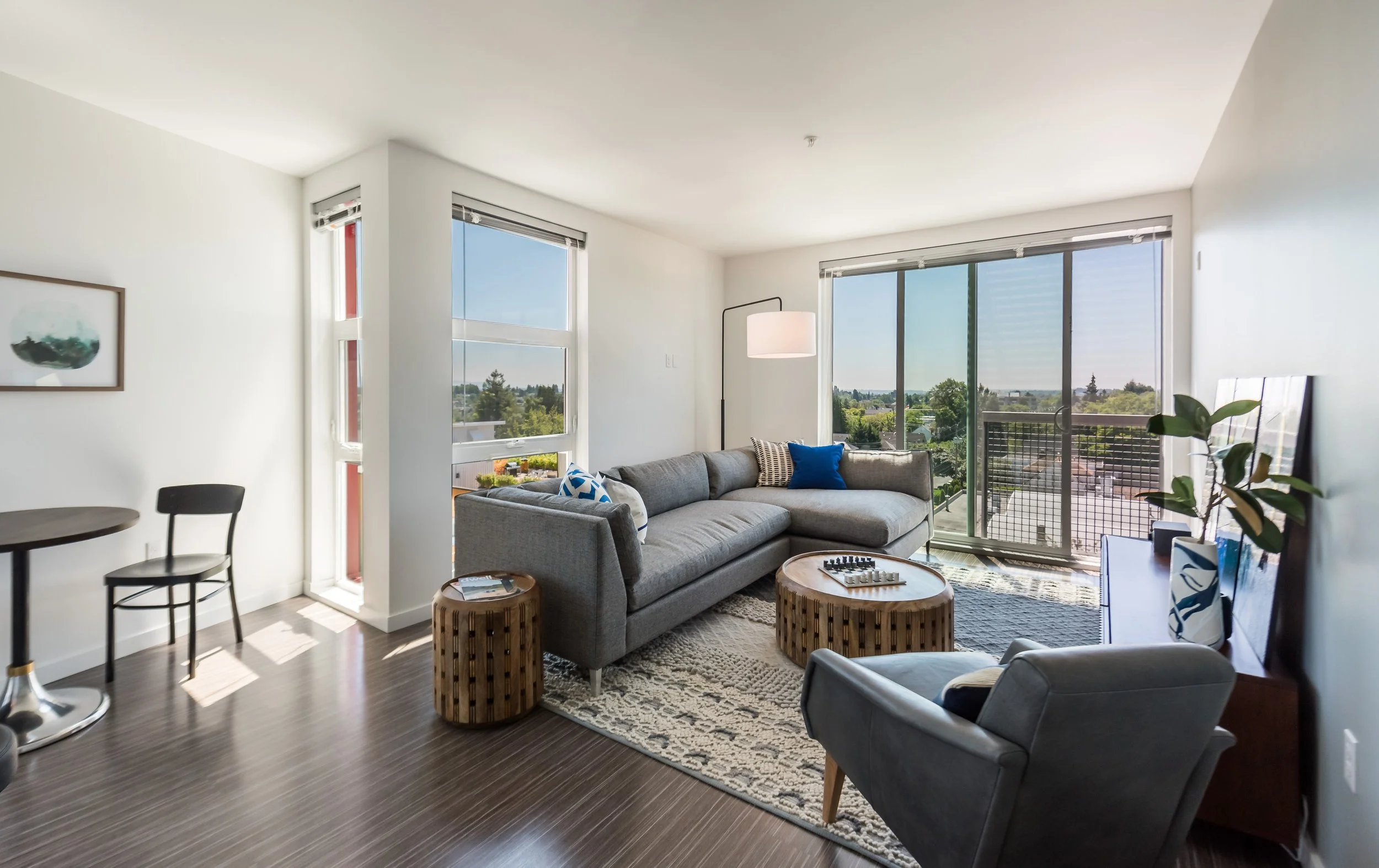 Living room with large windows, a gray sectional sofa with blue and patterned pillows, two round woven stools, a standing floor lamp, a chair, and a sliding glass door leading to a balcony with views of trees and buildings.