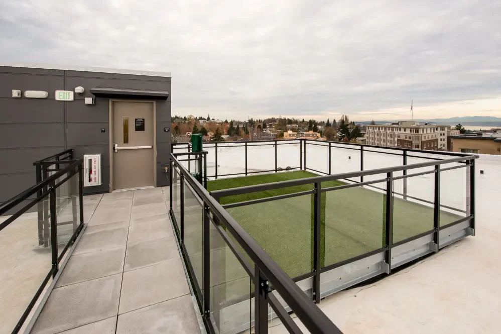Rooftop terrace with a small artificial grass area enclosed by glass panels, door, emergency fire box, and cityscape view in the background under cloudy sky.