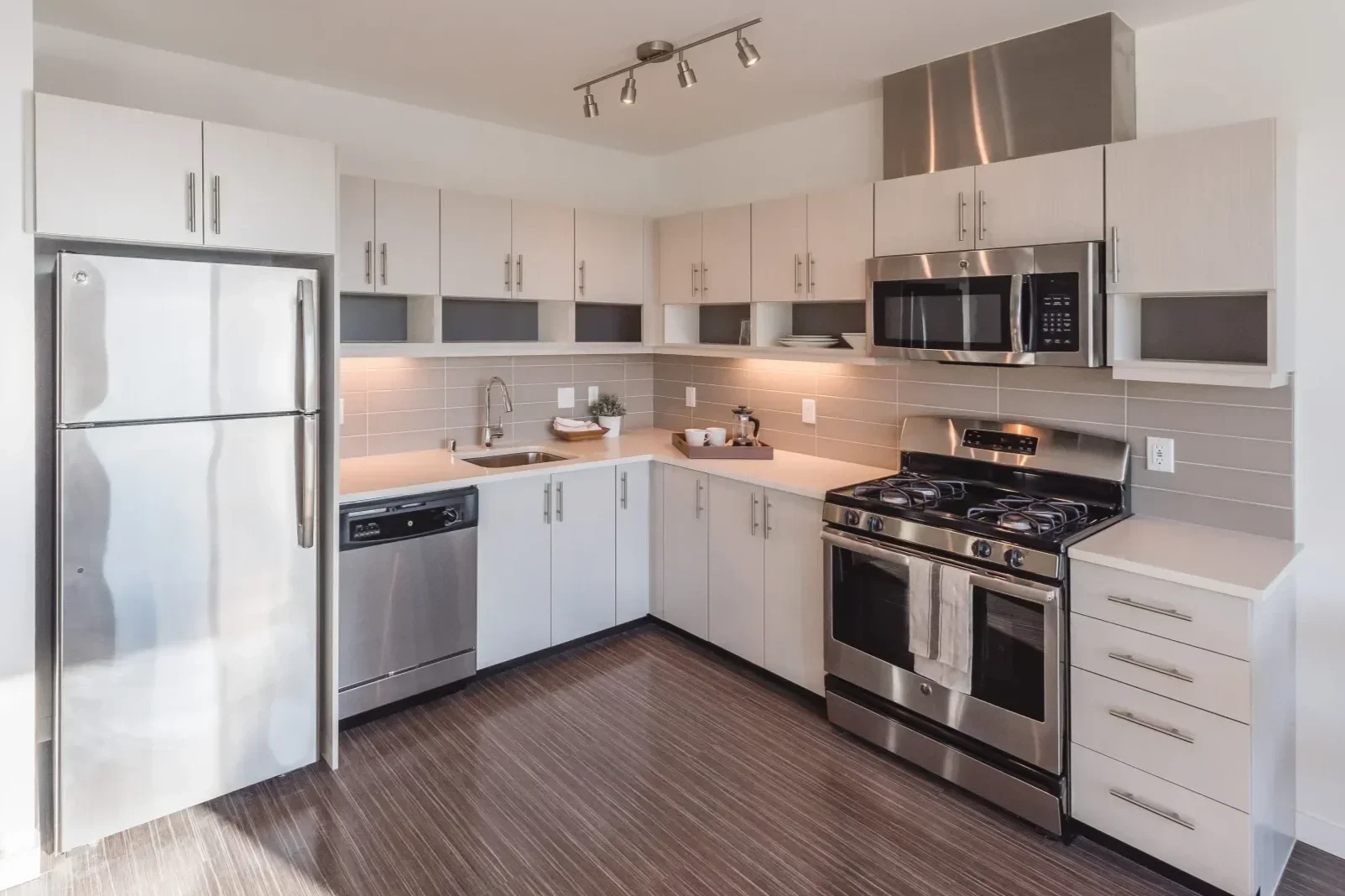 Modern kitchen with stainless steel appliances including a refrigerator, microwave, oven, and dishwasher, white cabinets, a gray tiled backsplash, a small sink, and wood-like flooring.