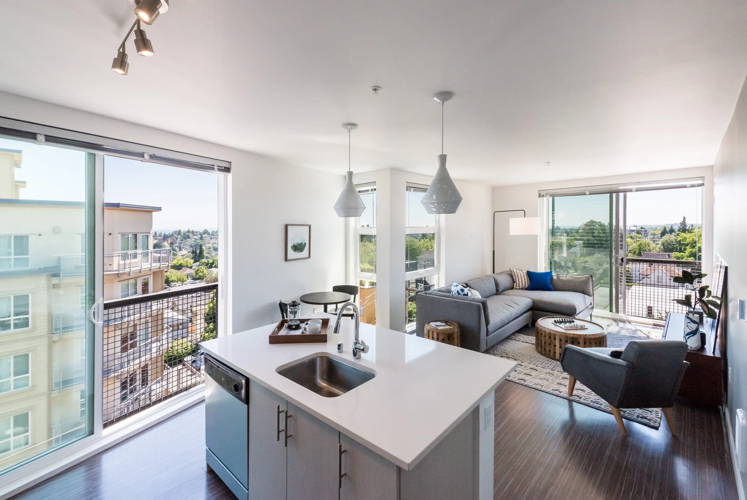 Interior of a modern apartment with an open floor plan, featuring a kitchen with a white island and a living area with a gray sofa, armchair, and large windows showing city views.