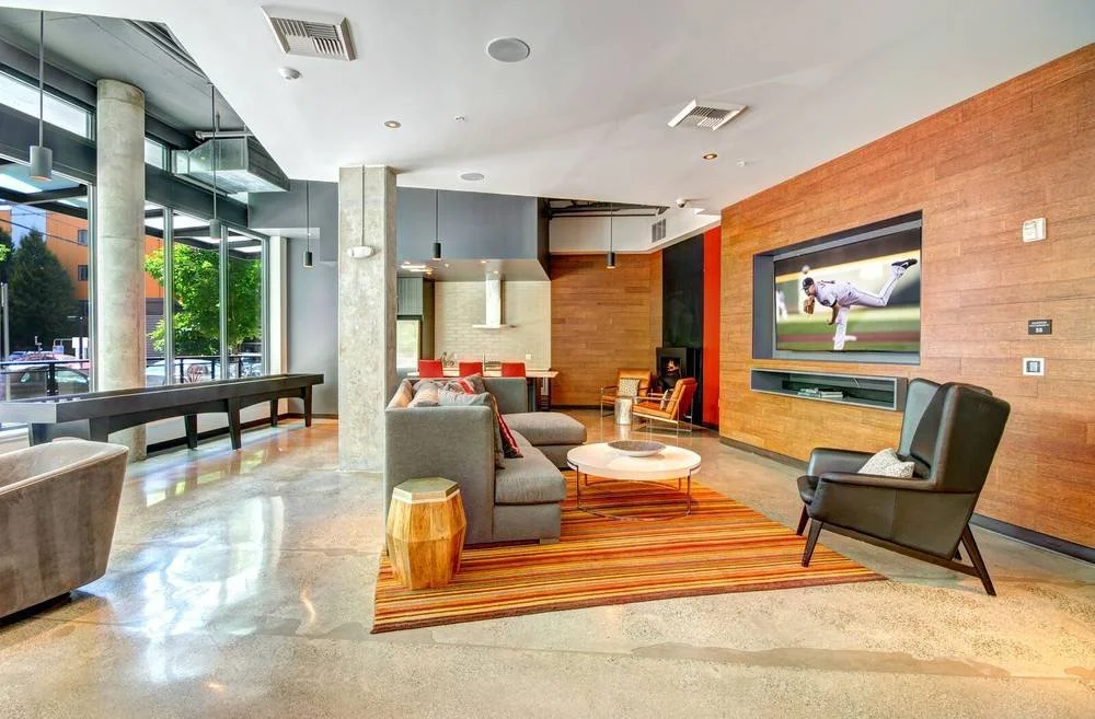 Modern hotel lobby with large windows, gray and beige furniture, a flat-screen TV on a wooden wall, and a striped rug.