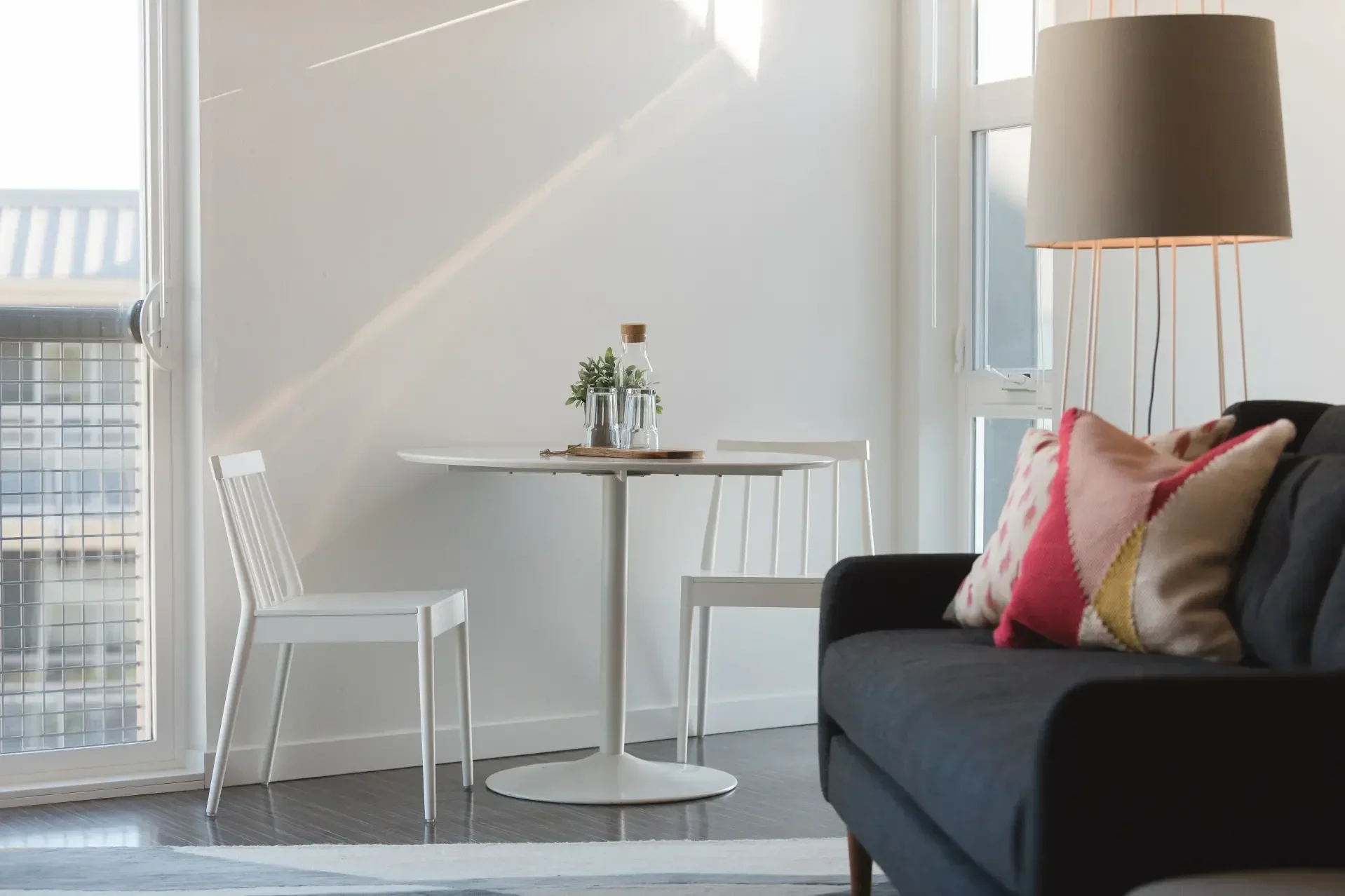 Living room with black sofa and pink, beige, and red patterned pillows. Small white round table near a white chair, decorated with a glass bottle, a small plant, and a tray. Large windows allow sunlight to fill the room. Floor has dark wood planks, a