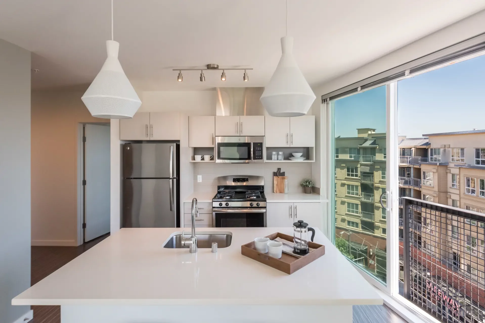 Modern kitchen with white cabinets, stainless steel appliances including a refrigerator, microwave, and stove, a white countertop island with a sink, and large windows showing neighboring buildings and a balcony.
