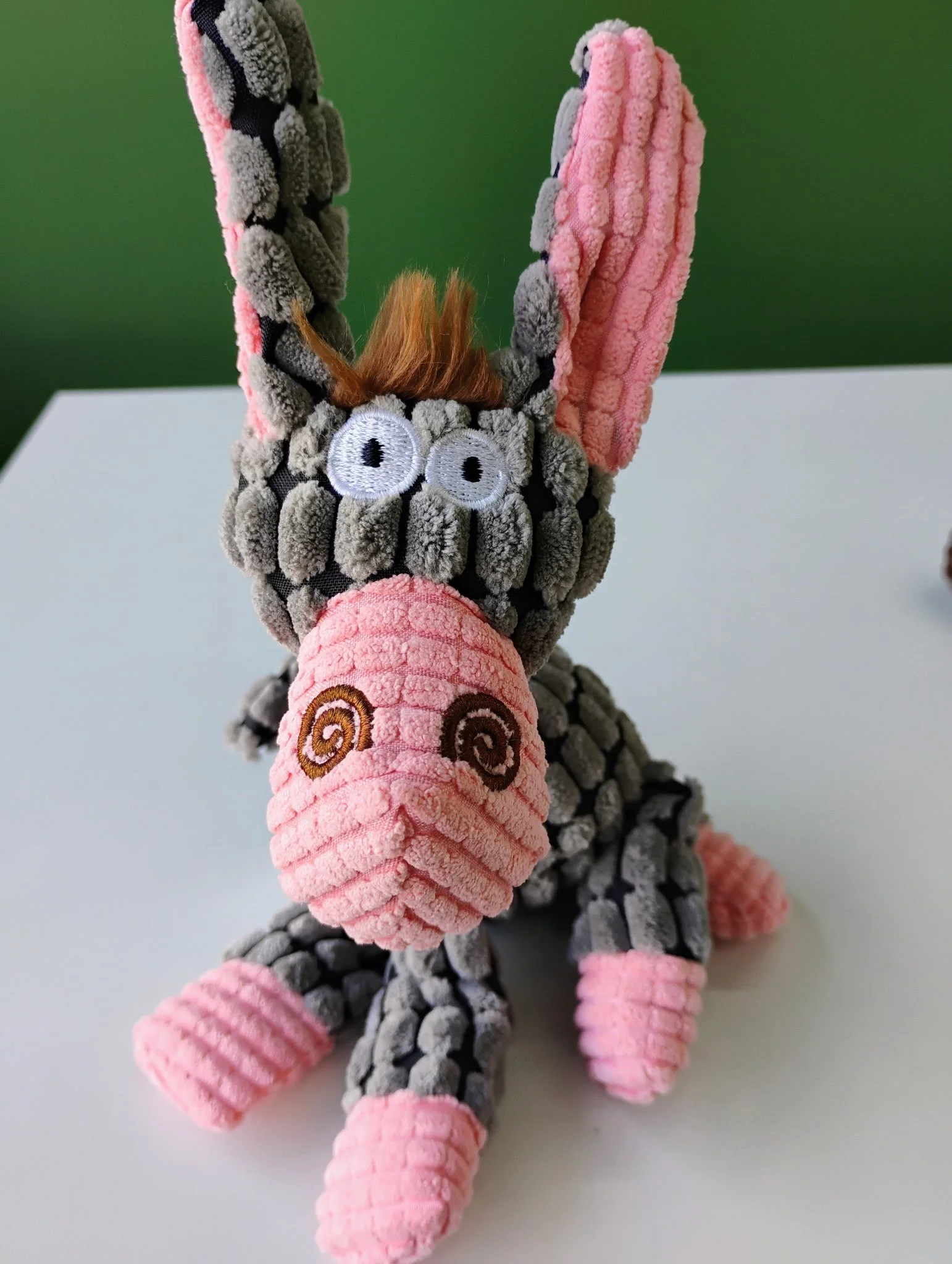 A plush toy resembling a kangaroo made of soft, textured fabric with pink, gray, black, and white colors. The toy has embroidered eyes, a small nose, and a pink pouch with spiral embroidery, standing against a green background.