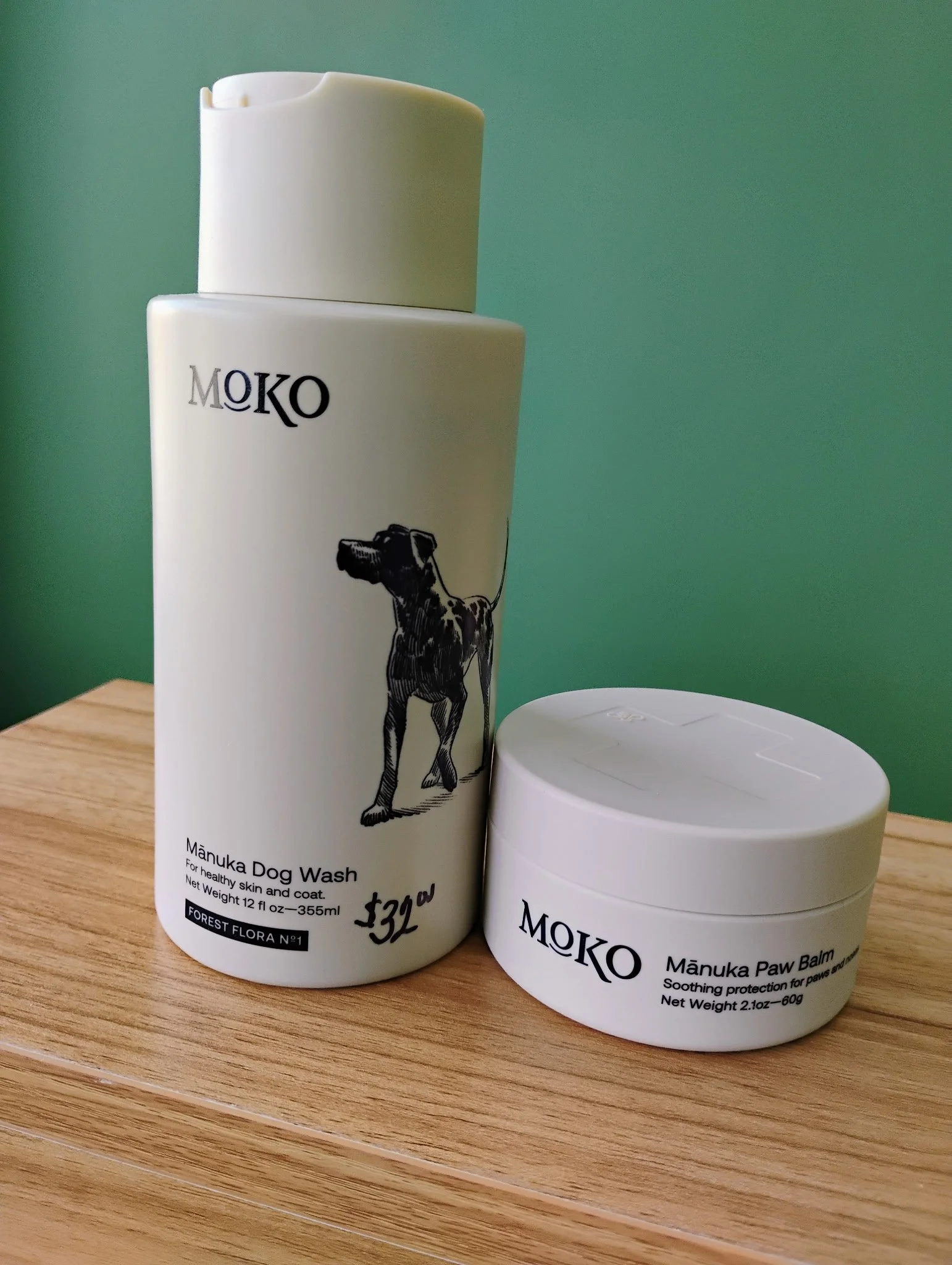 A white bottle of MŌKO Manuka Dog Wash and a white container of MŌKO Manuka Paw Balm on a wooden surface with a green background.