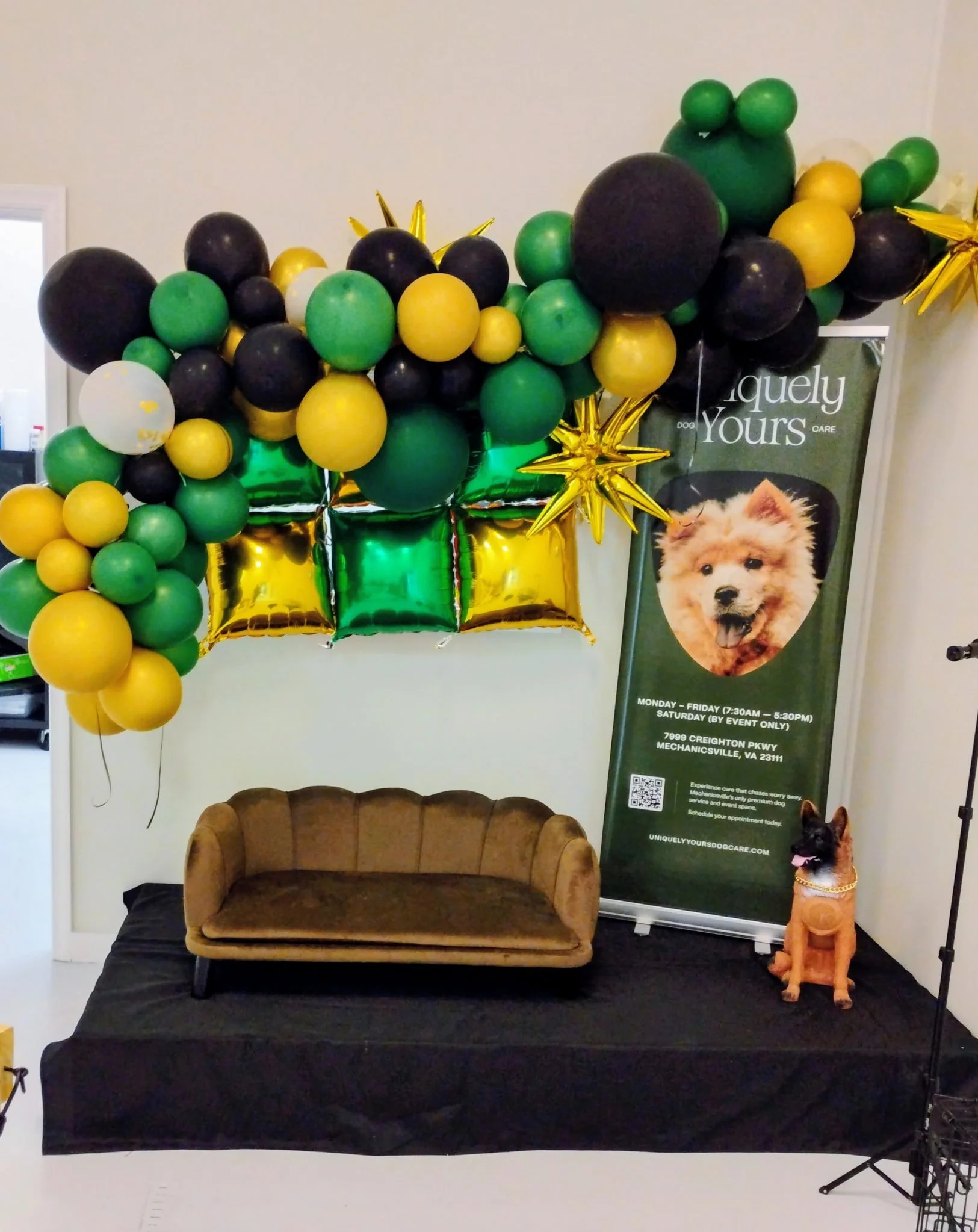 Decorative setup including a tan velvet sofa on a black tablecloth, green and yellow balloons, gold star-shaped balloons, a green and white poster advertising dog grooming services, and a life-sized stuffed dog figure with a gold collar.