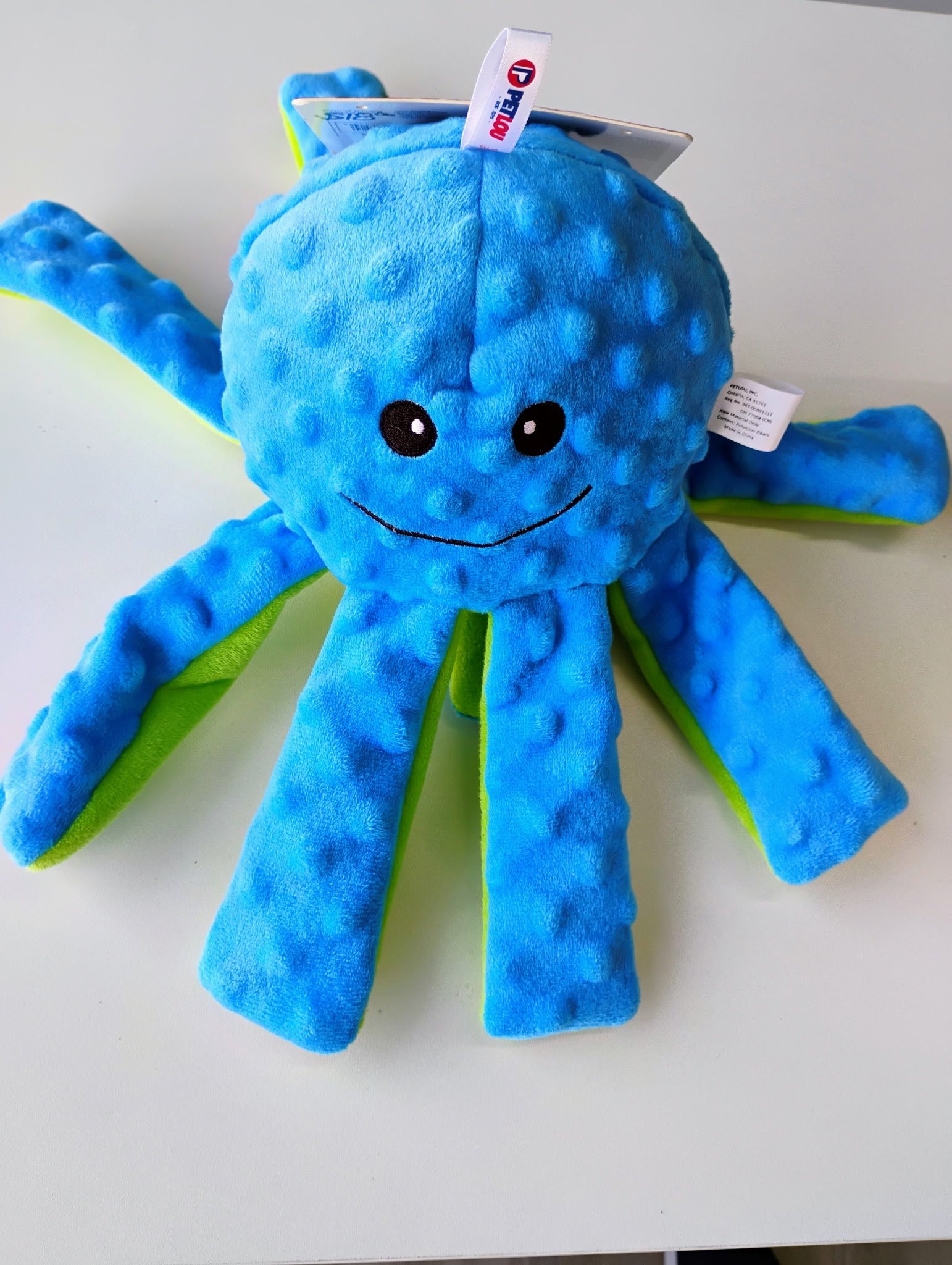 A blue plush octopus toy with black embroidered eyes and a smile, with yellow and green accents on its arms, on a white surface.