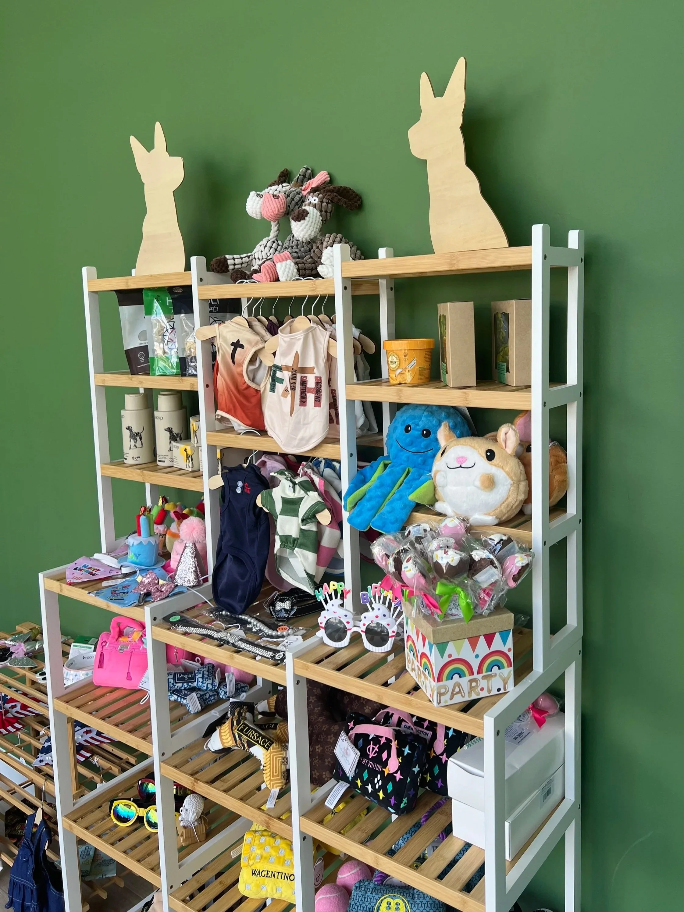 Display of various accessories and toys on a multi-tiered wooden and white metal shelf against a green wall. Items include plush toys, sunglasses, children's clothes, bags, and party favors.