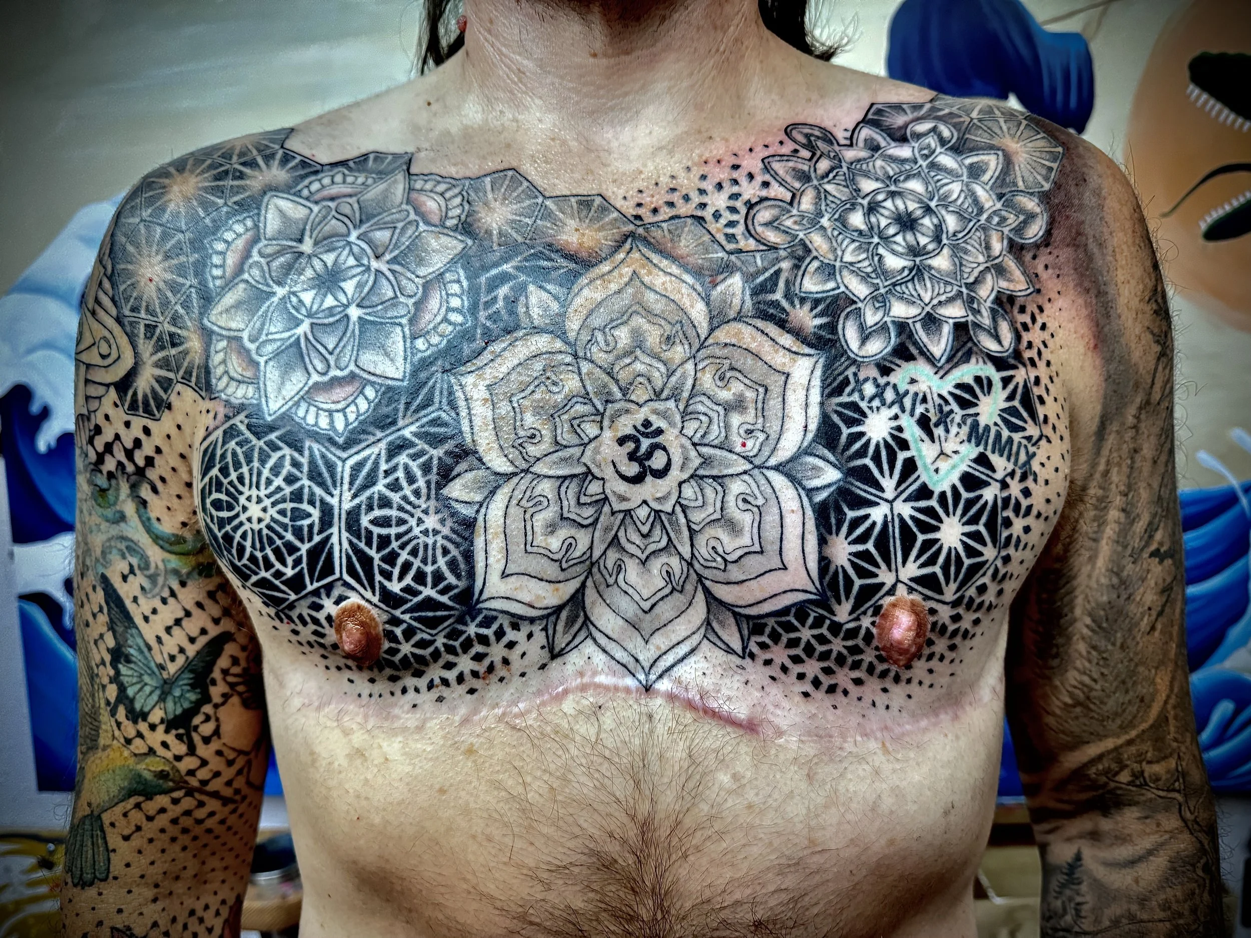 A person with tattoos on their chest, shoulders, and arms, including mandala designs, a lotus flower with an 'Om' symbol, and geometric patterns, undergoing a tattoo session.