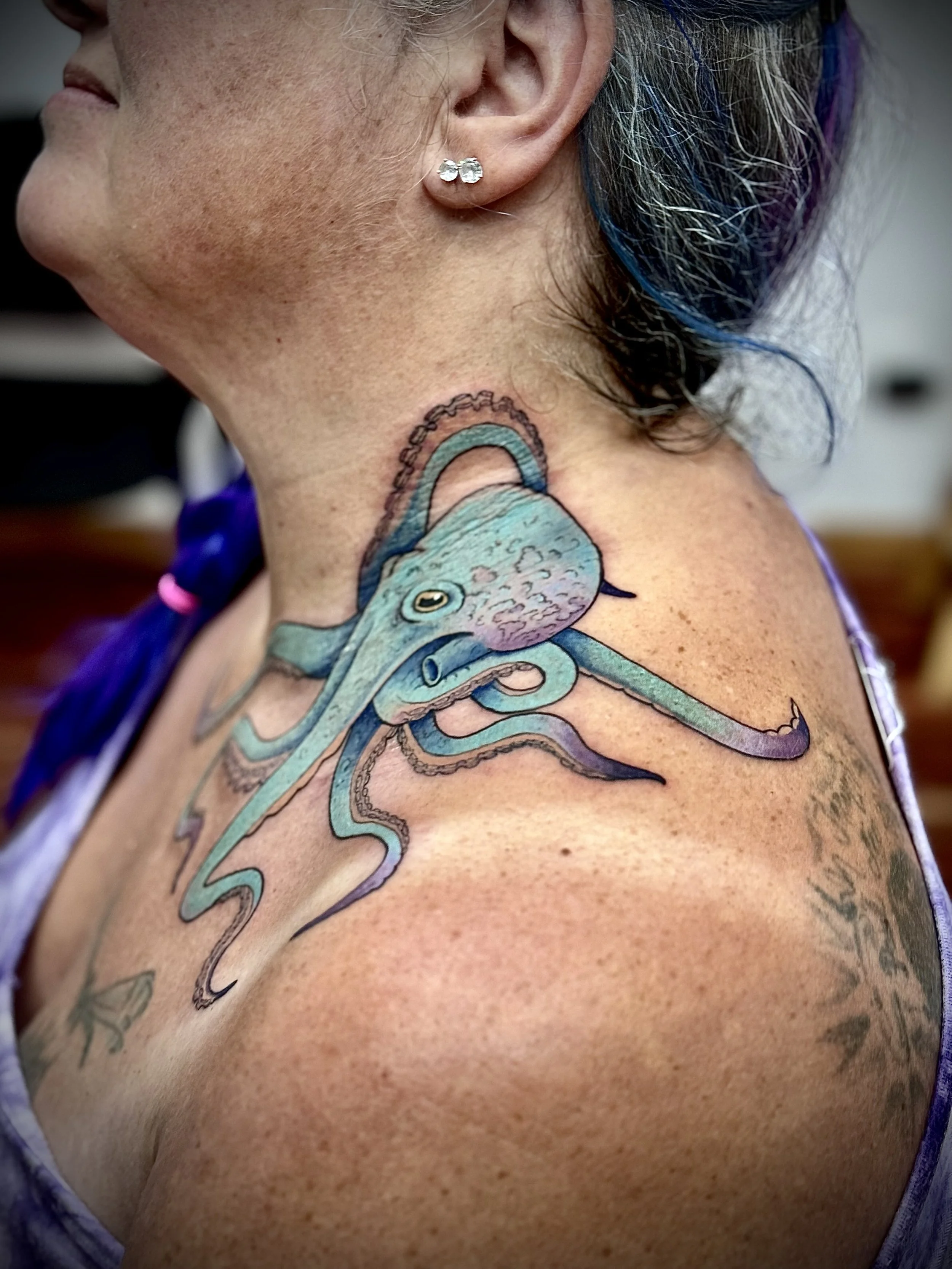 Close-up of a woman's shoulder and neck showing a colorful octopus tattoo with detailed shading, along with her ear and part of her face, with rainbow-colored hair strands visible.