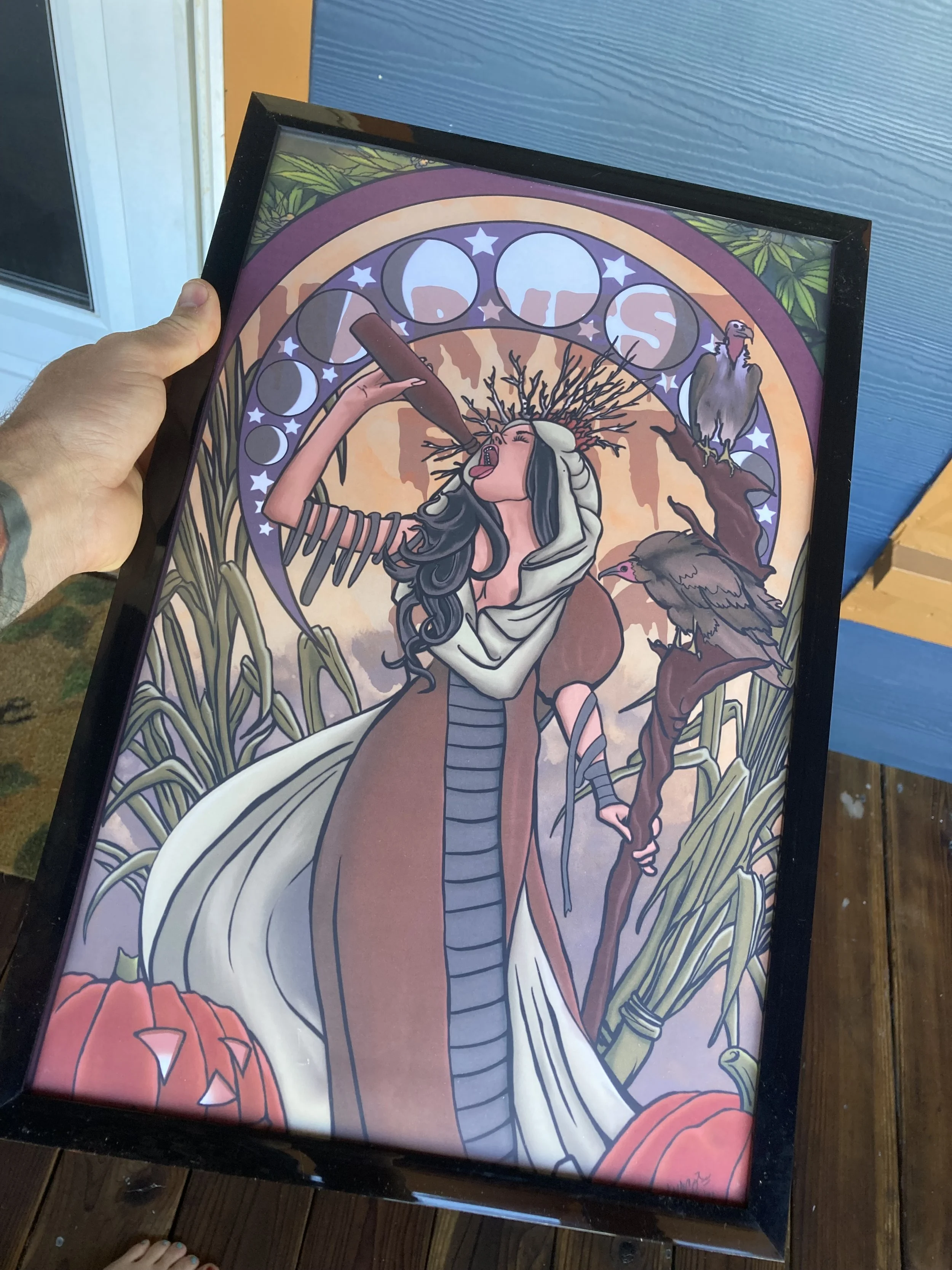 Framed illustration of a woman with long dark hair, wearing a white head covering and white robes, holding a sickle in her right hand, standing amidst tall grasses and pumpkins. Behind her is a stained-glass-like circular design with stars and moon phases. Two turkeys are on her left and right, and a barnyard scene is visible at the bottom.