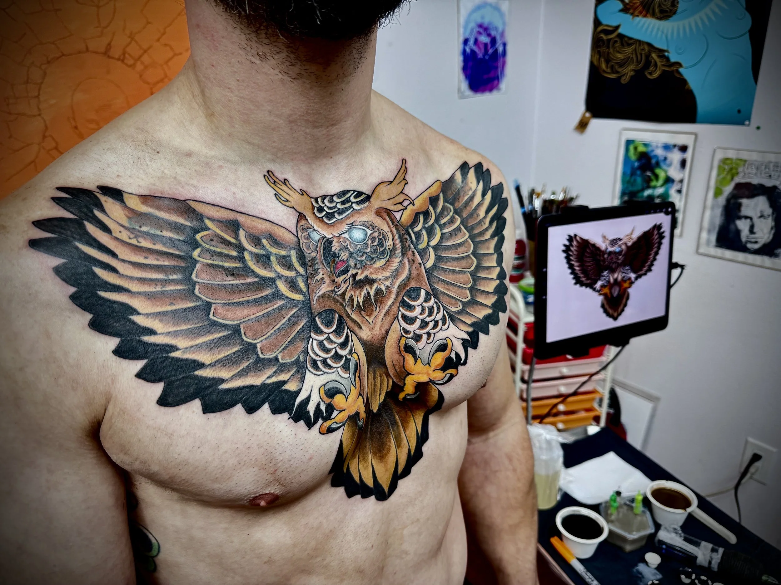 A person with a tattoo of a colorful owl on their chest, showing details of the owl's feathers and face, in a tattoo studio with artwork and tattoo supplies in the background.