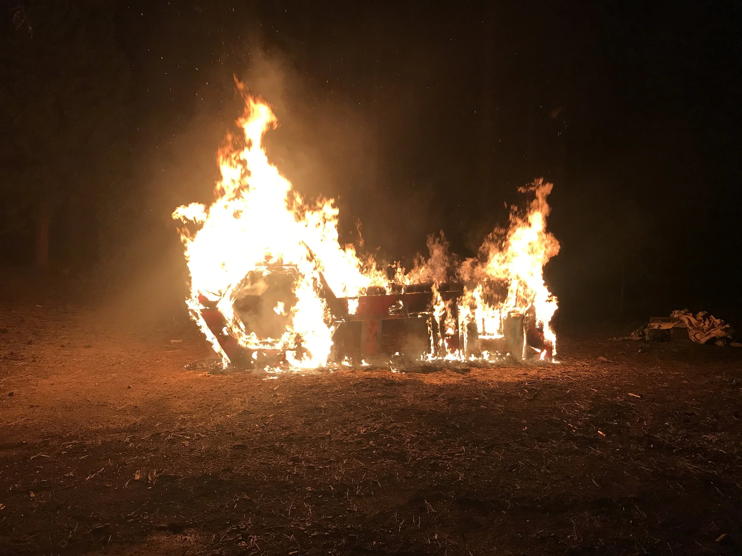 Bonfire with flames and sparks rising into the night sky, situated on a patch of dirt or grass, with a dark, starry sky in the background.