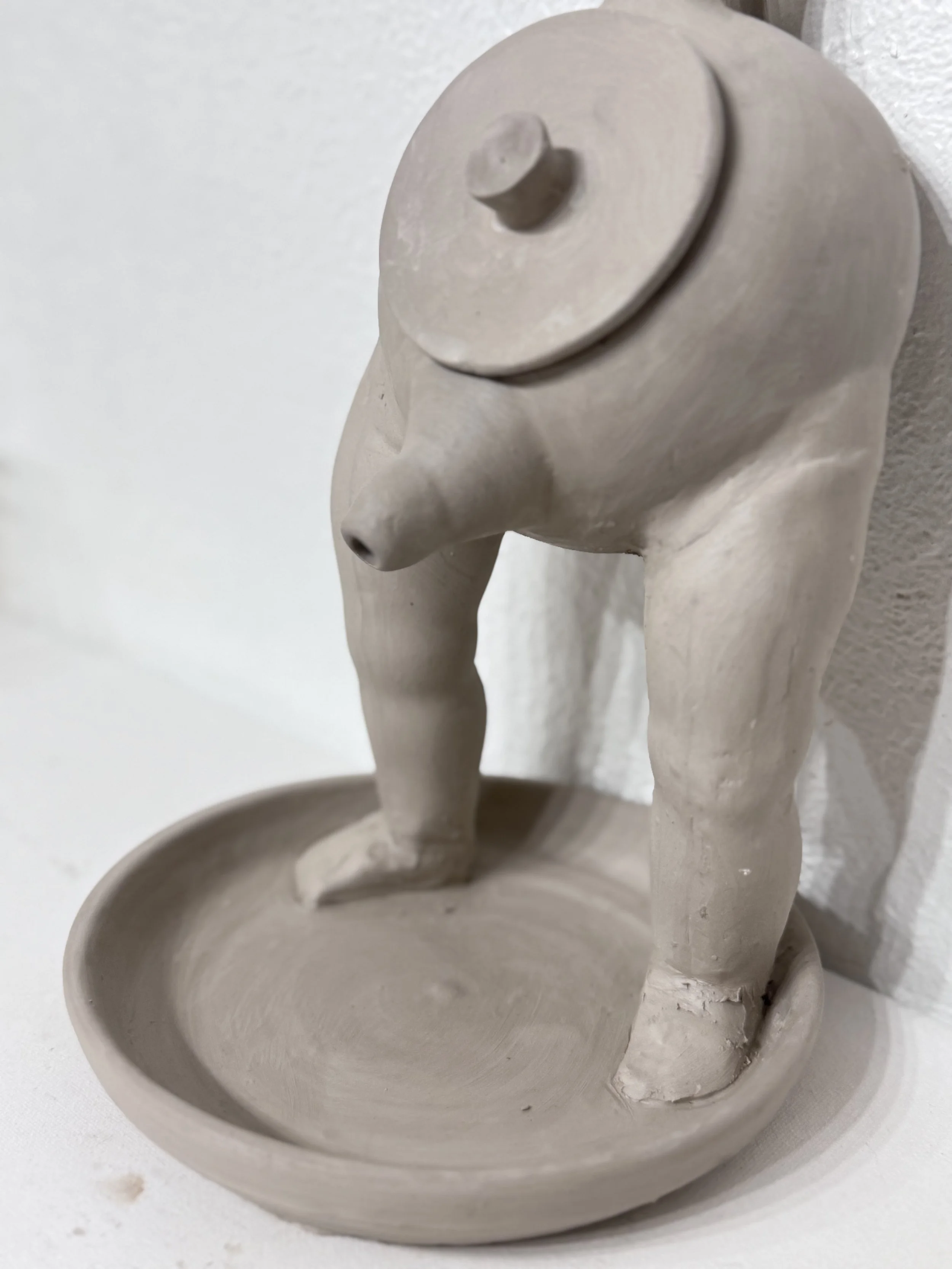 A small, abstract ceramic sculpture resembling a human figure with a large, rounded head and elongated body, positioned over a shallow, bowl-like base.