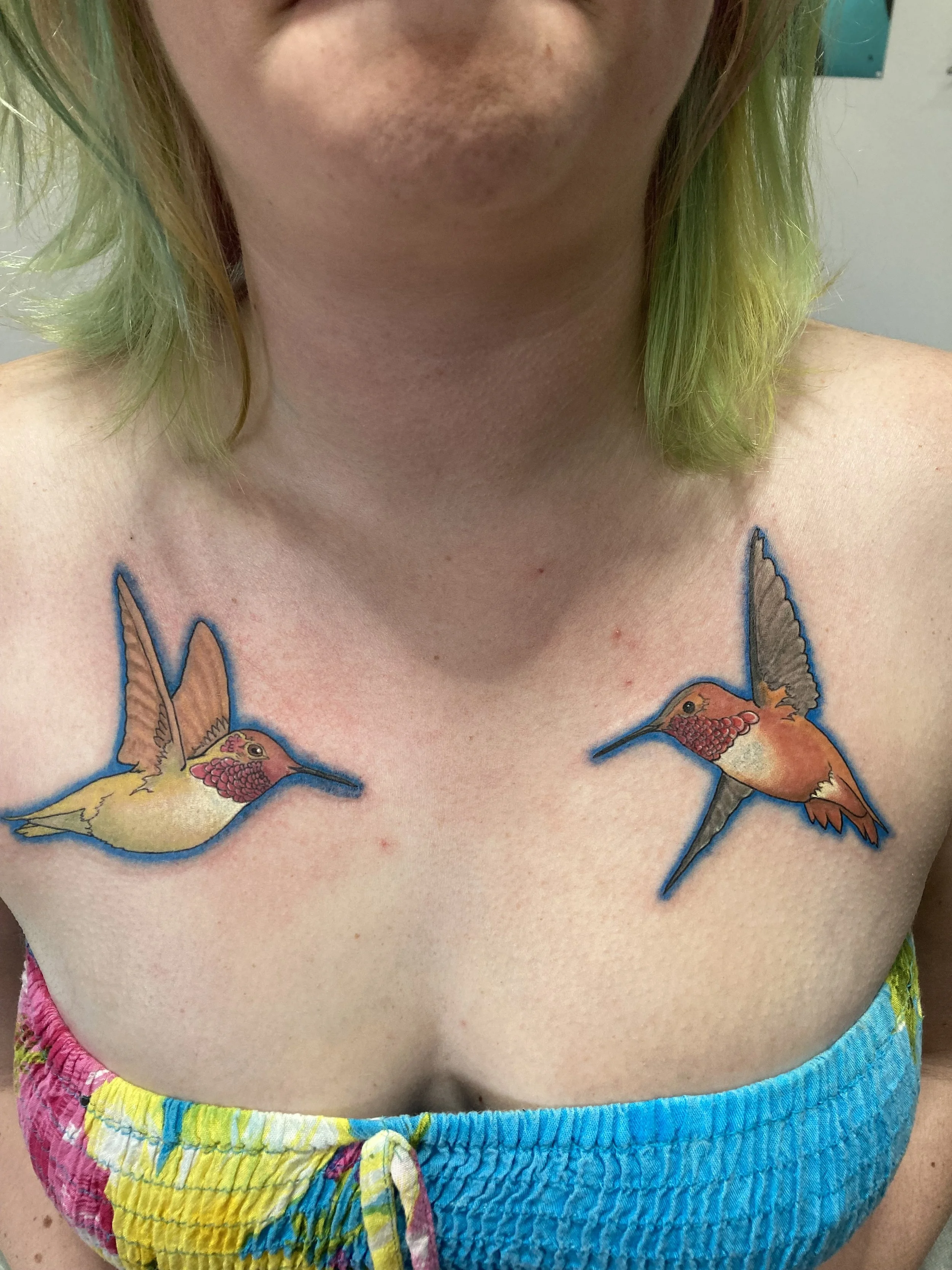 Close-up of a person's chest with a tattoo of two colorful hummingbirds.