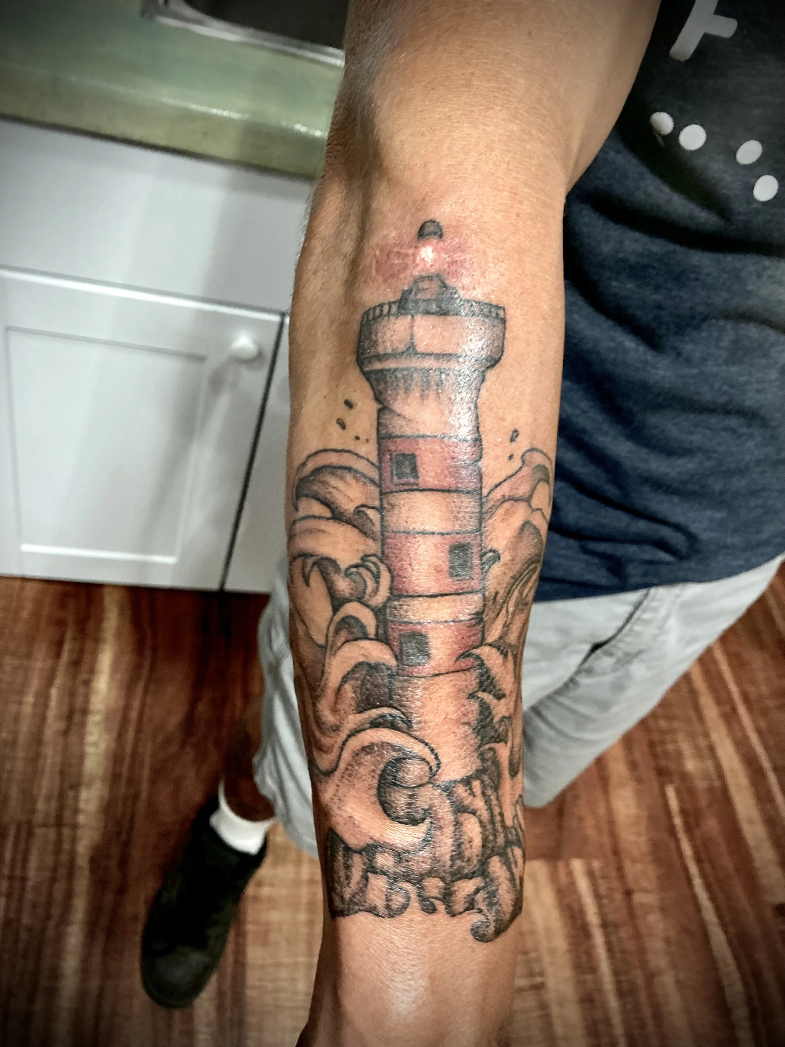 Close-up of a person's forearm with a tattoo of a lighthouse and text, with a background of wooden flooring and part of a person's leg wearing gray pants and black shoes.
