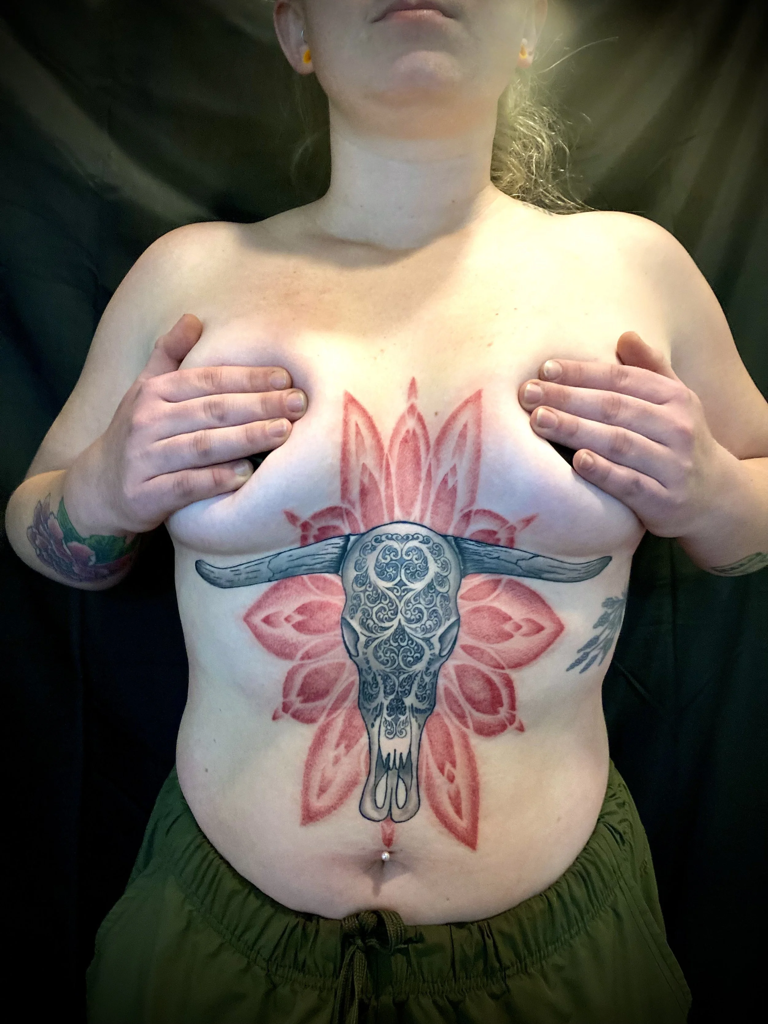 A woman sleeping on her back with her hands covering her breasts. She has large tattoos on her chest and arms, and a decorative tattoo of a longhorn skull with intricate blue designs on her abdomen. She is wearing green pants and earrings, and has light-colored hair.