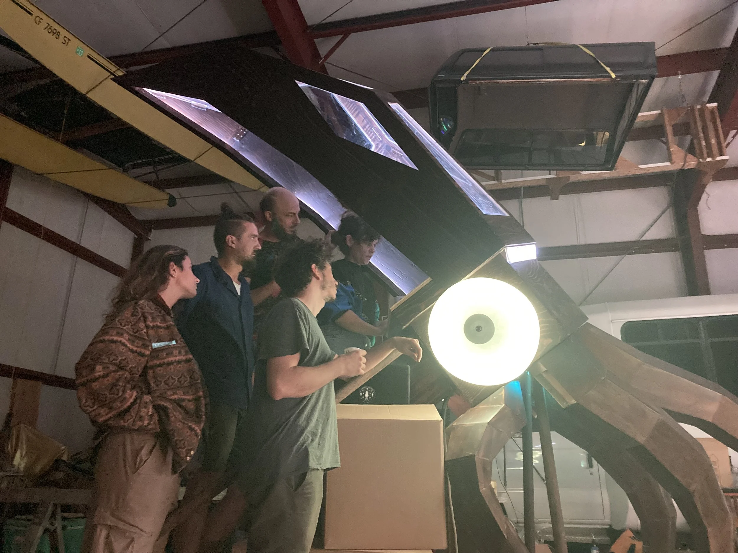 Group of people standing and reading in a dimly lit indoor space, next to large illuminated spherical objects and scattered furniture.