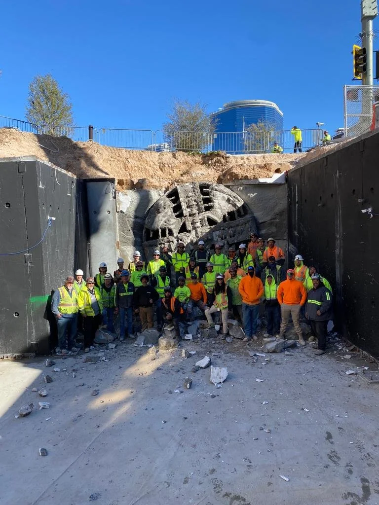 Group of construction workers and engineers posing in front of a tunnel excavation site with a tunnel entrance and construction equipment, under a clear blue sky.
