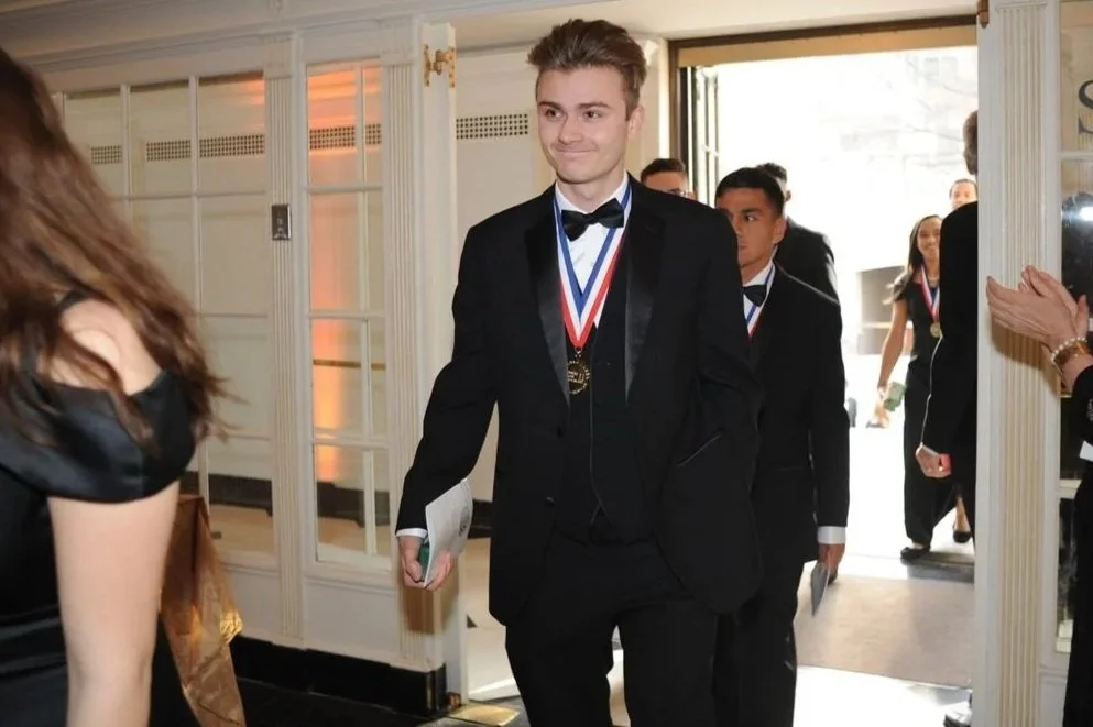 A young man in a black tuxedo with a bow tie and a gold medal around his neck walking inside a building. Other people in tuxedos are visible in the background, some wearing medals. A woman with long brown hair in a black dress is on the left side of the image.