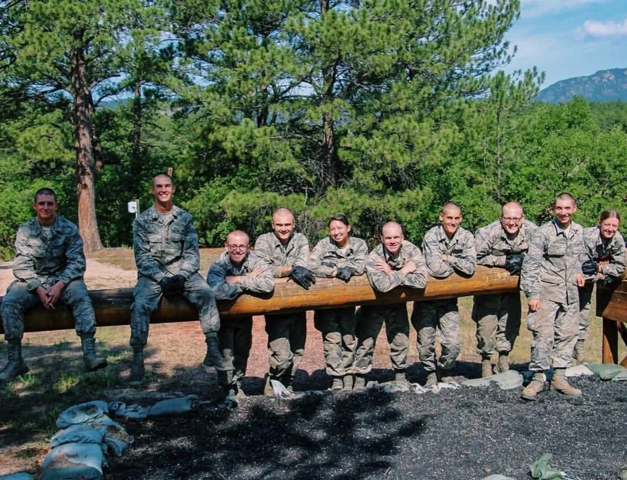 A group of ten soldiers in military uniforms are leaning on a large wooden log outdoors with trees and mountains in the background.
