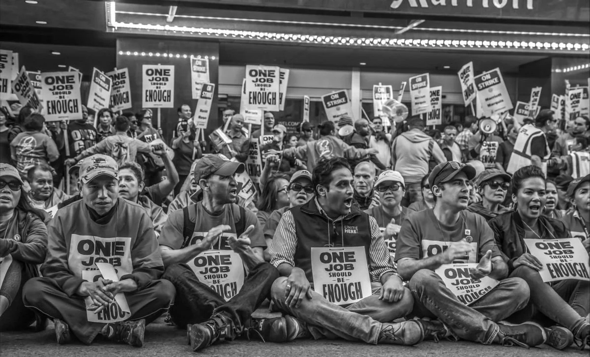 Crowd of protesters sitting on the floor holding signs that read "One Job Should Be Enough" during a demonstration.