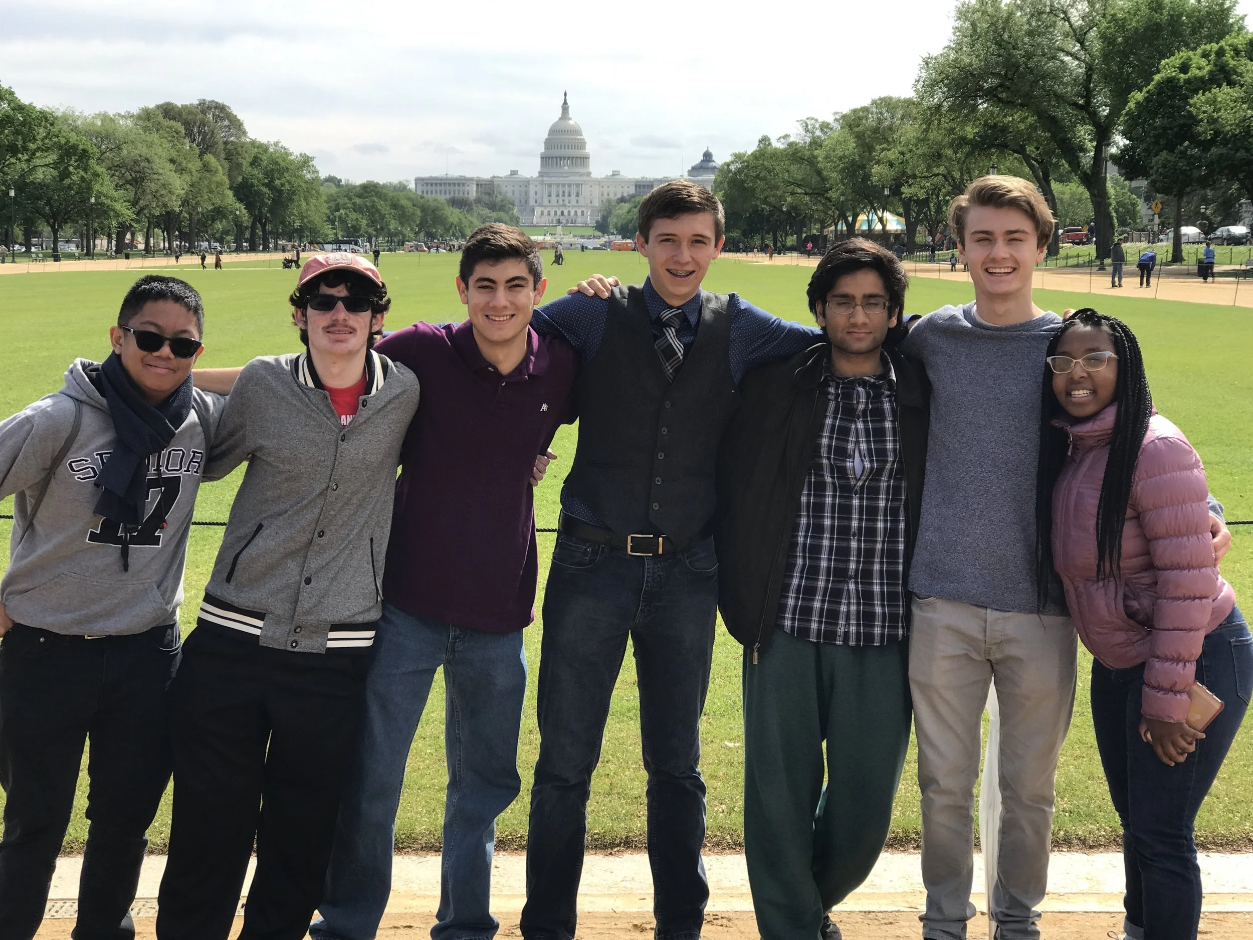 Group of eight teenagers standing arm-in-arm in front of the U.S. Capitol building in Washington, D.C., smiling at the camera on a partly cloudy day.