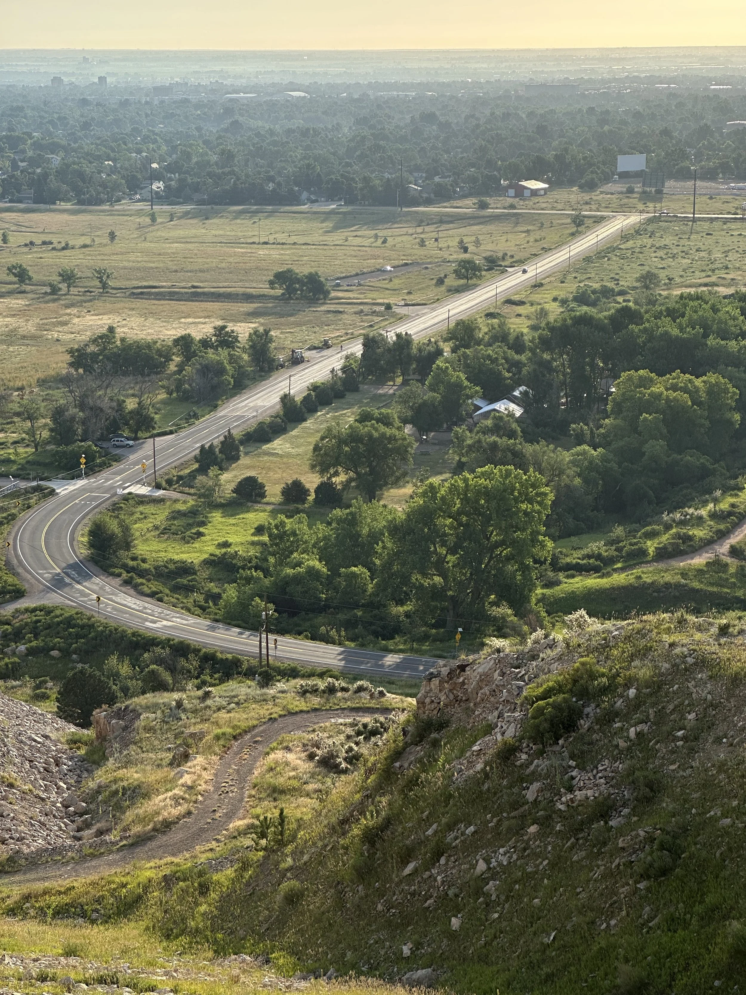 Aerial view of a winding road through green countryside, with trees, fields, and distant city skyline on the horizon.